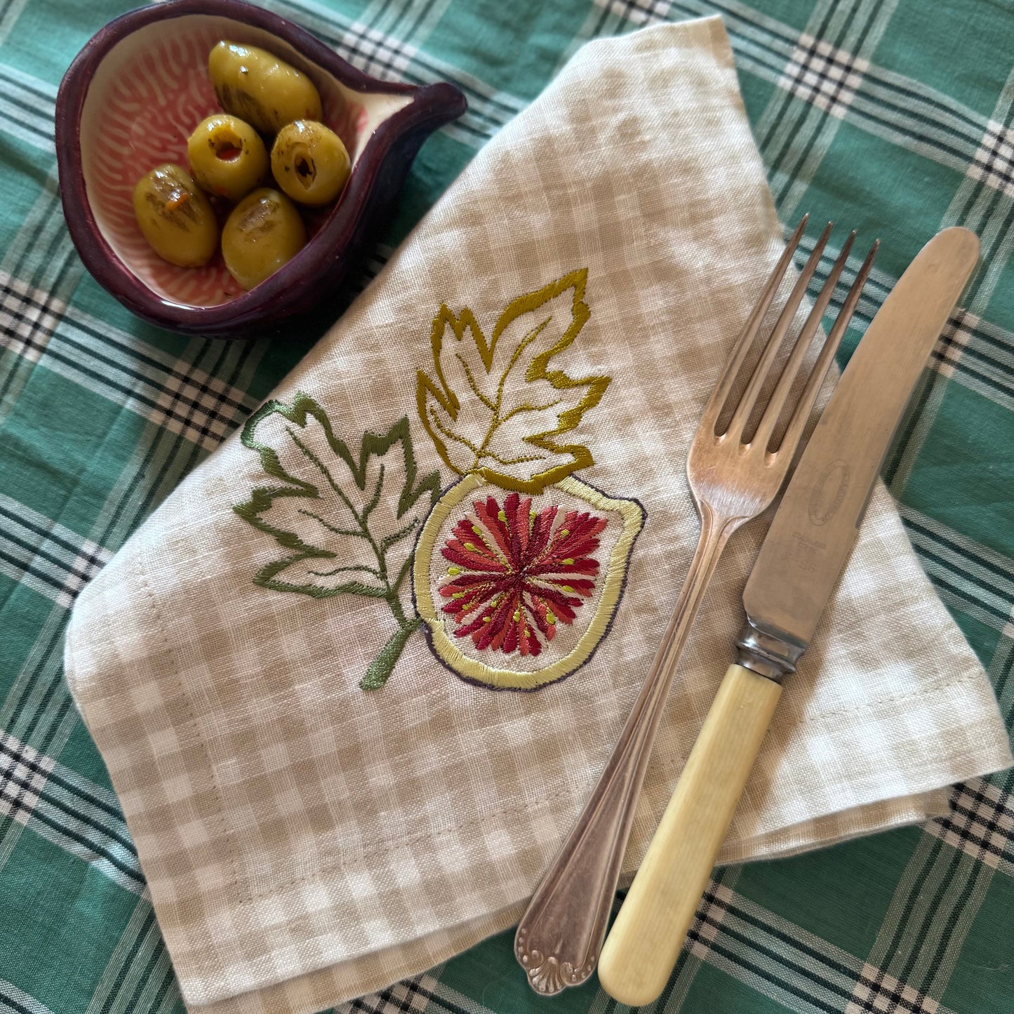 Napkin with fig design, fork, and knife on a checkered tablecloth with olives.