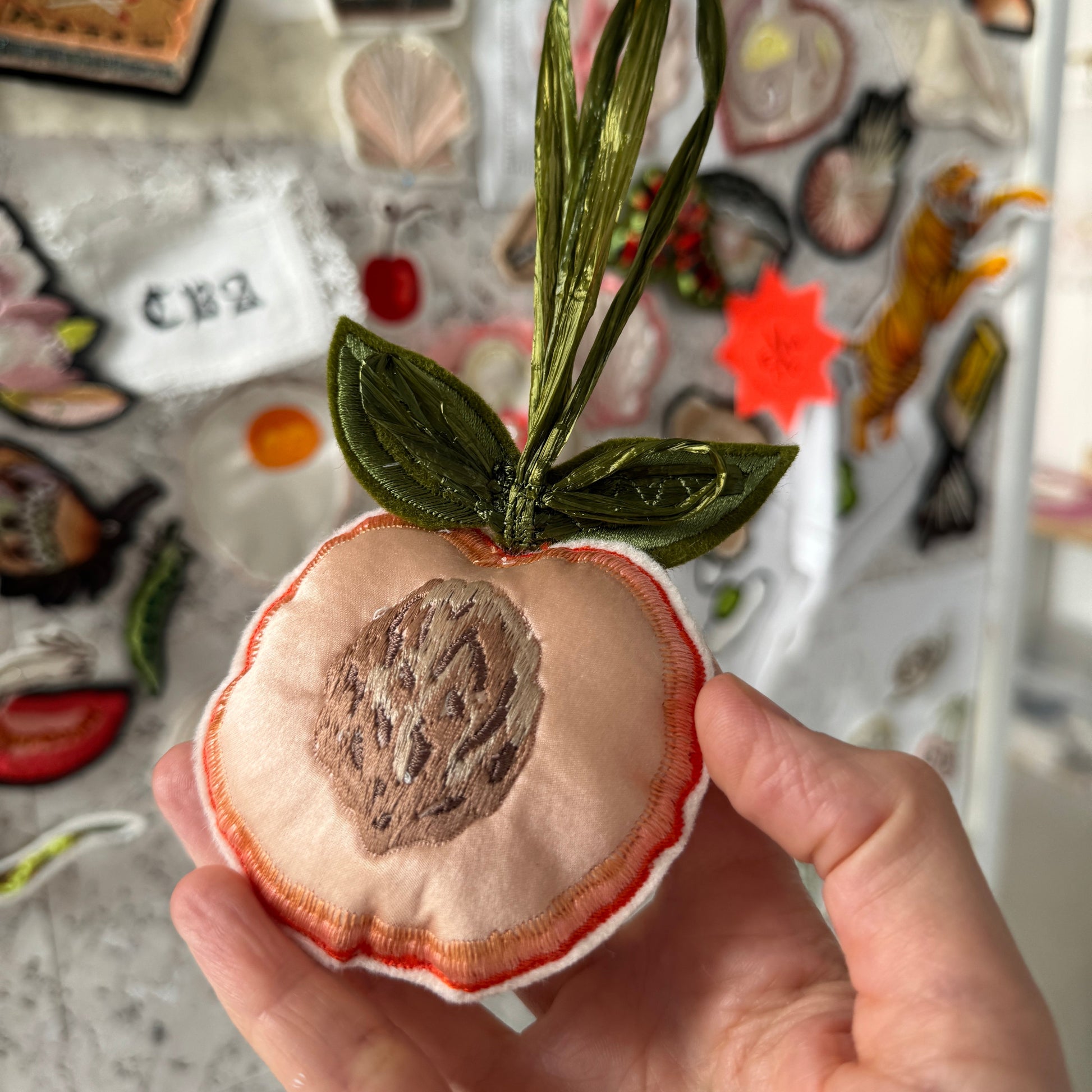 Hand holding a small fabric peach with green leaves against a colorful background