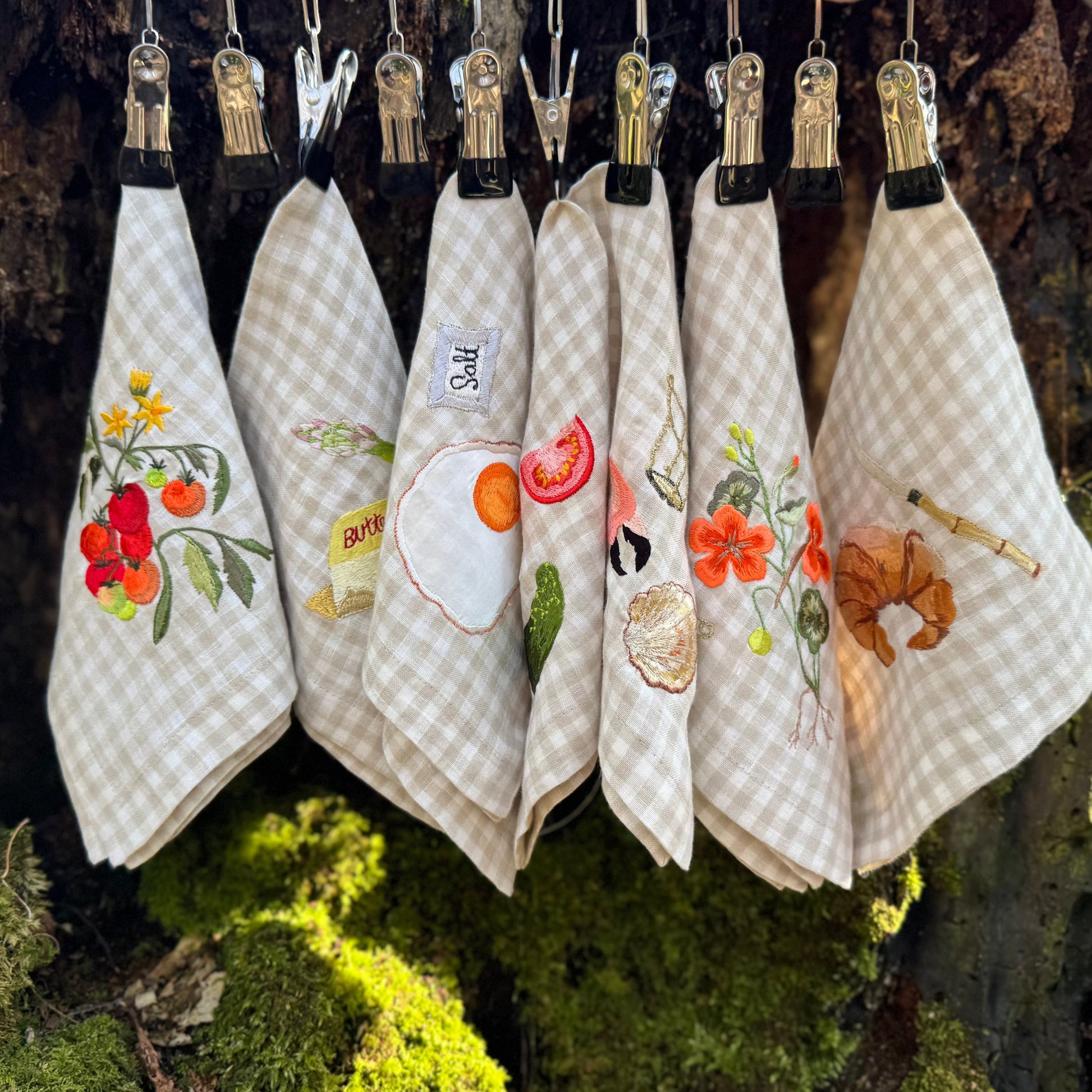 Collection of embroidered linen napkins on small steel pegs hanging in front of a mossy tree