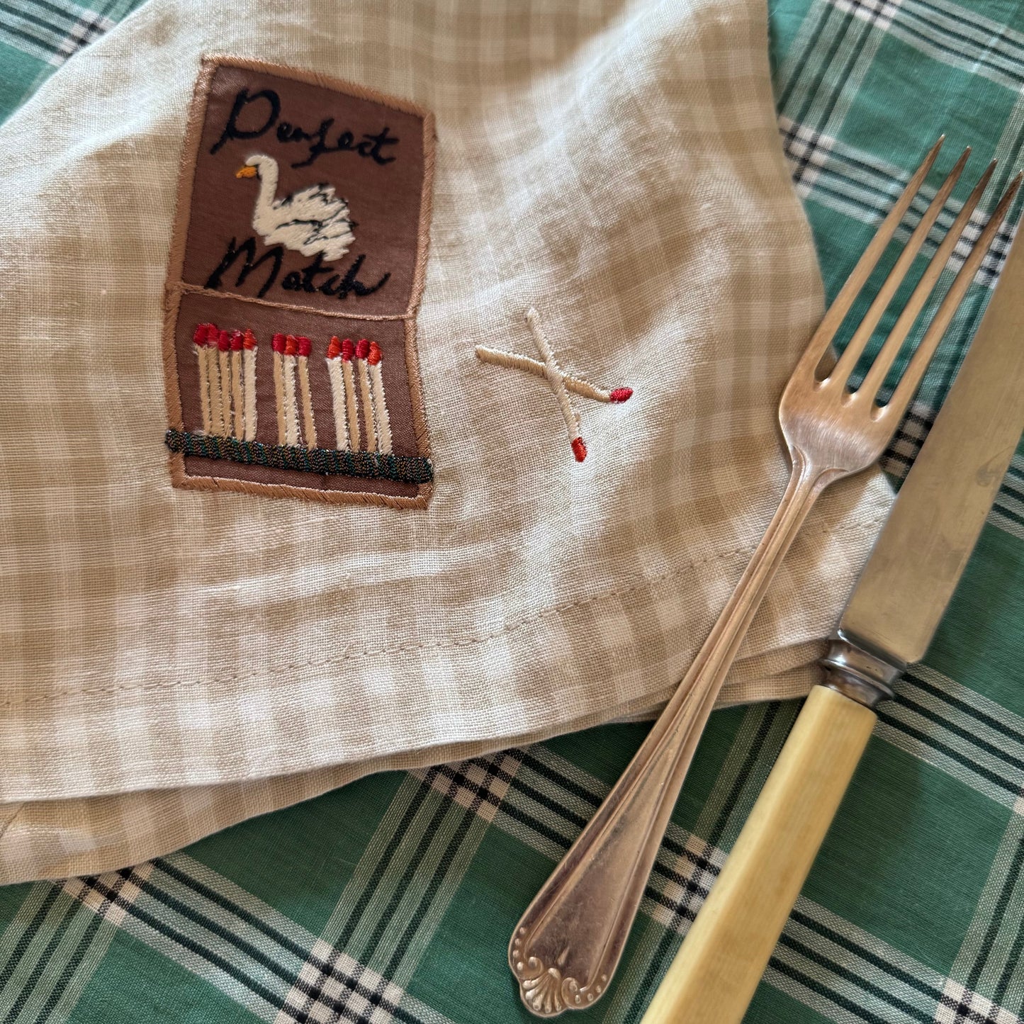 Napkin with embroidered design of a matchbox and matchstick, placed on a green checkered tablecloth with a fork and knife.