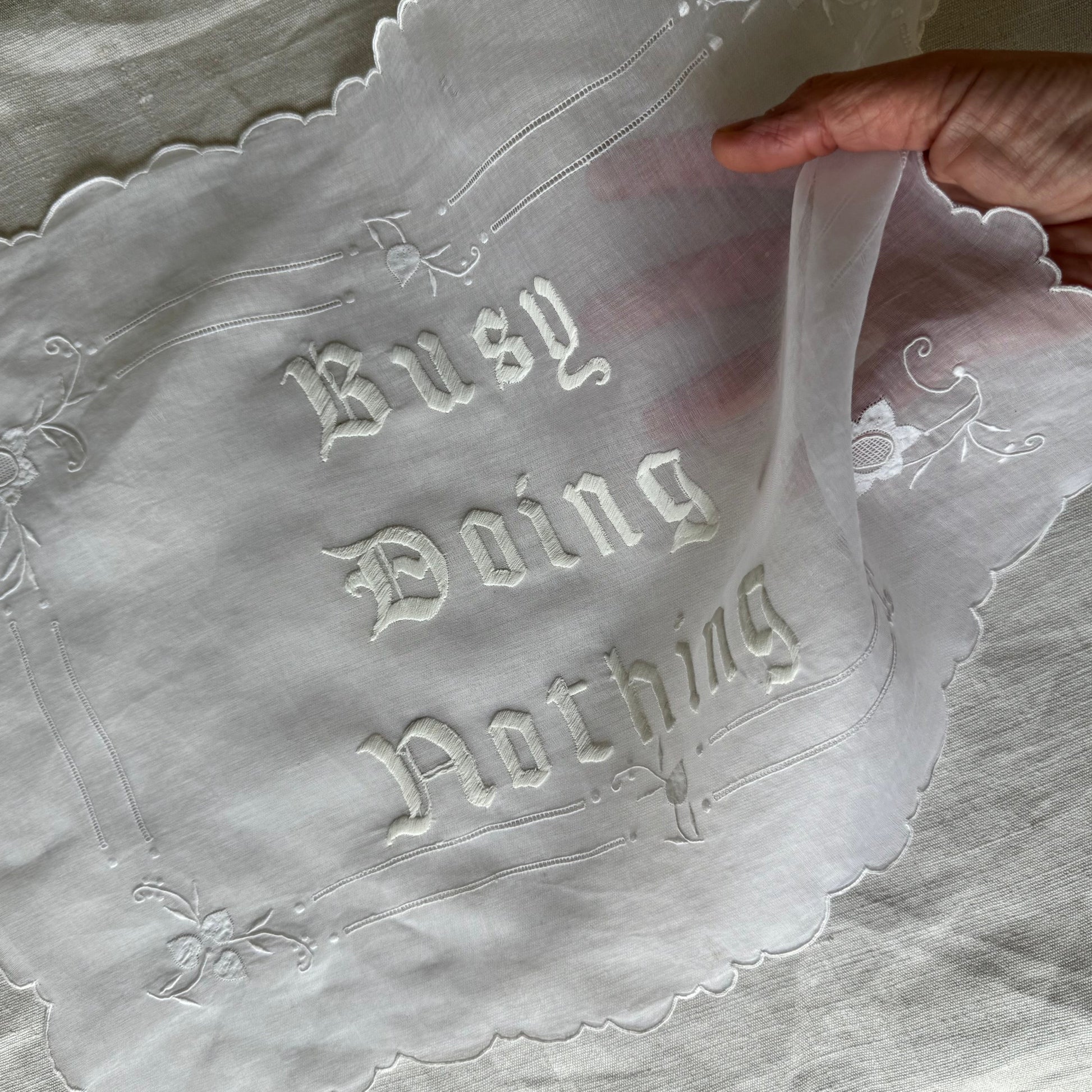 Busy Doing Nothing embroidered artwork on a piece of white fabric and a hand is seen holding one corner of the artwork
