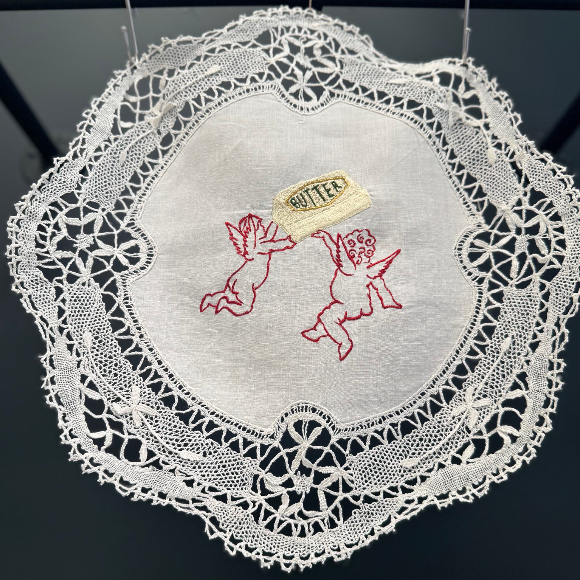 Bespoke butter babies linen artwork order on a vintage lace doiley. Photographed hanging in front of a dark grey wall