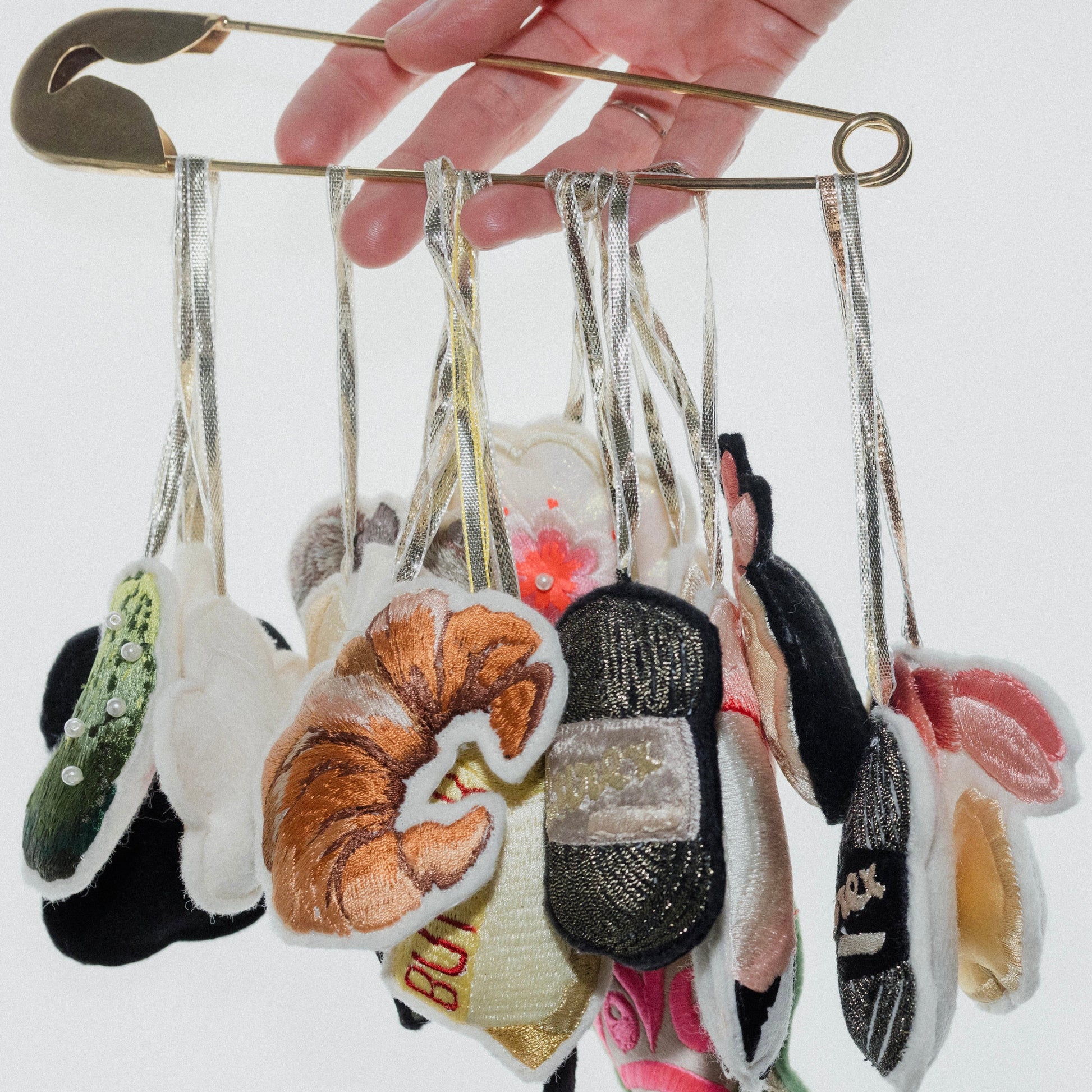 Hand holding a safety pin with various embroidered hanging decorations.