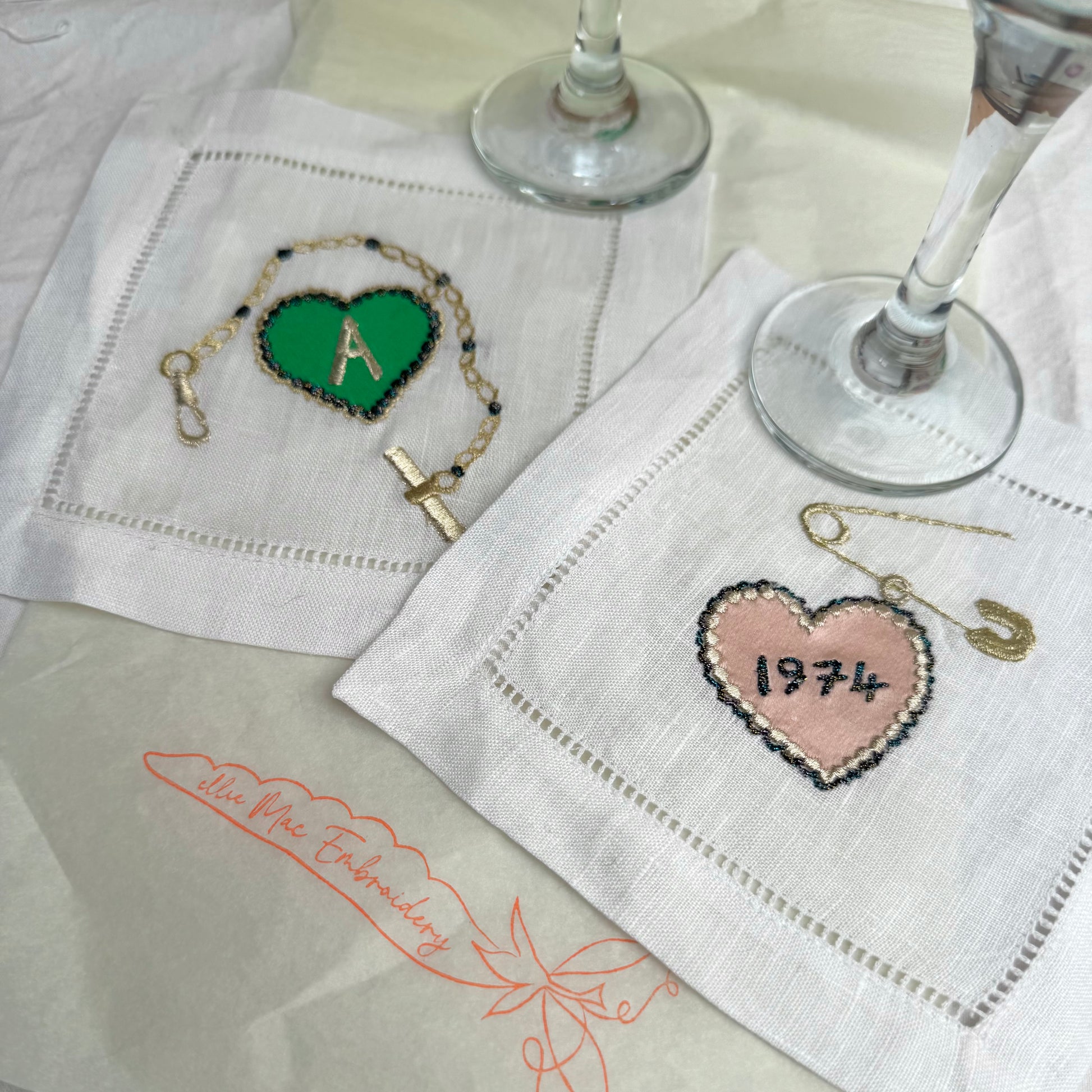 Heart charm and gold bracelet charm embroidered artworks on a table with the bases of a pair of wine glasses just visible