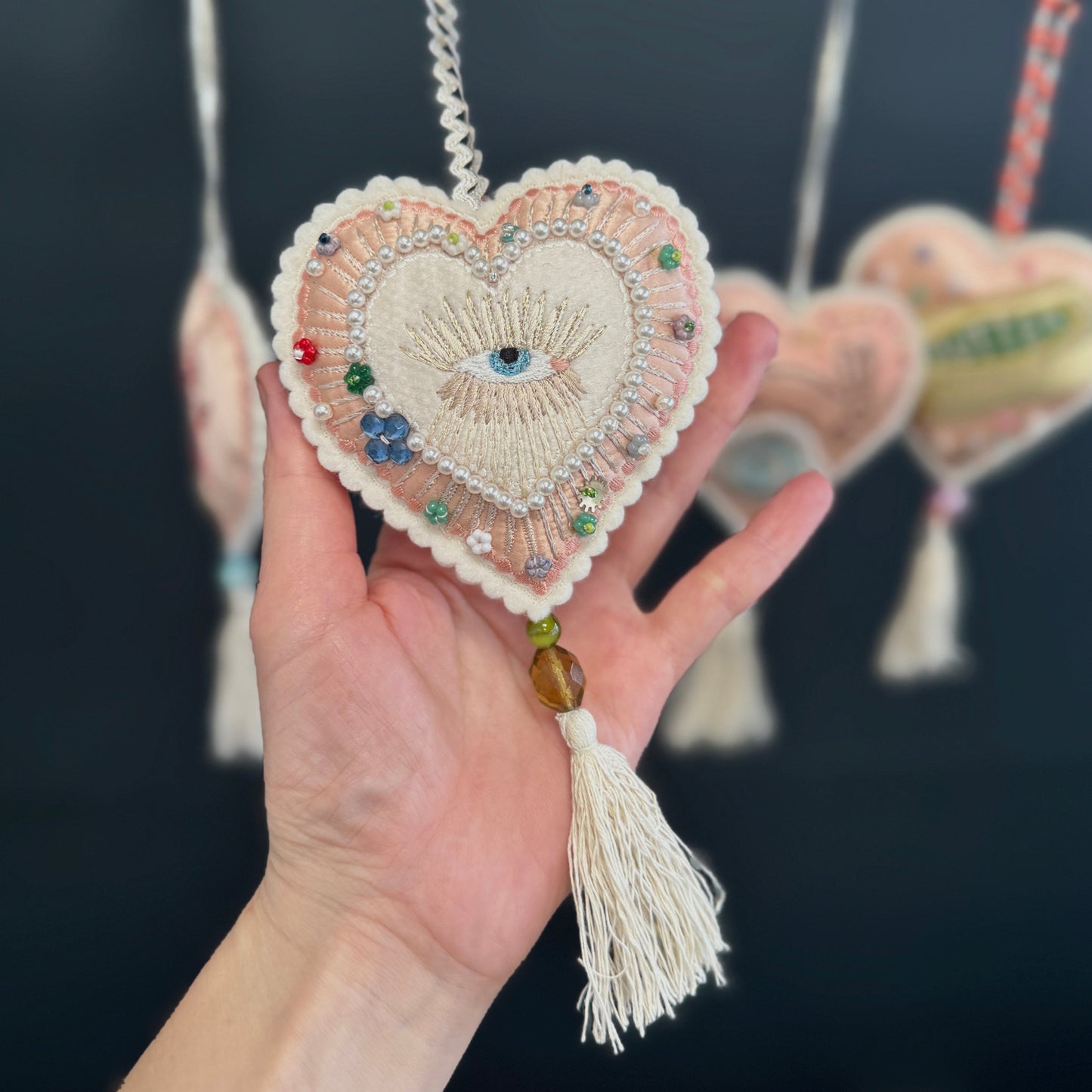 Heart-shaped embroidered ornament with beads and tassel held in a hand against a dark background