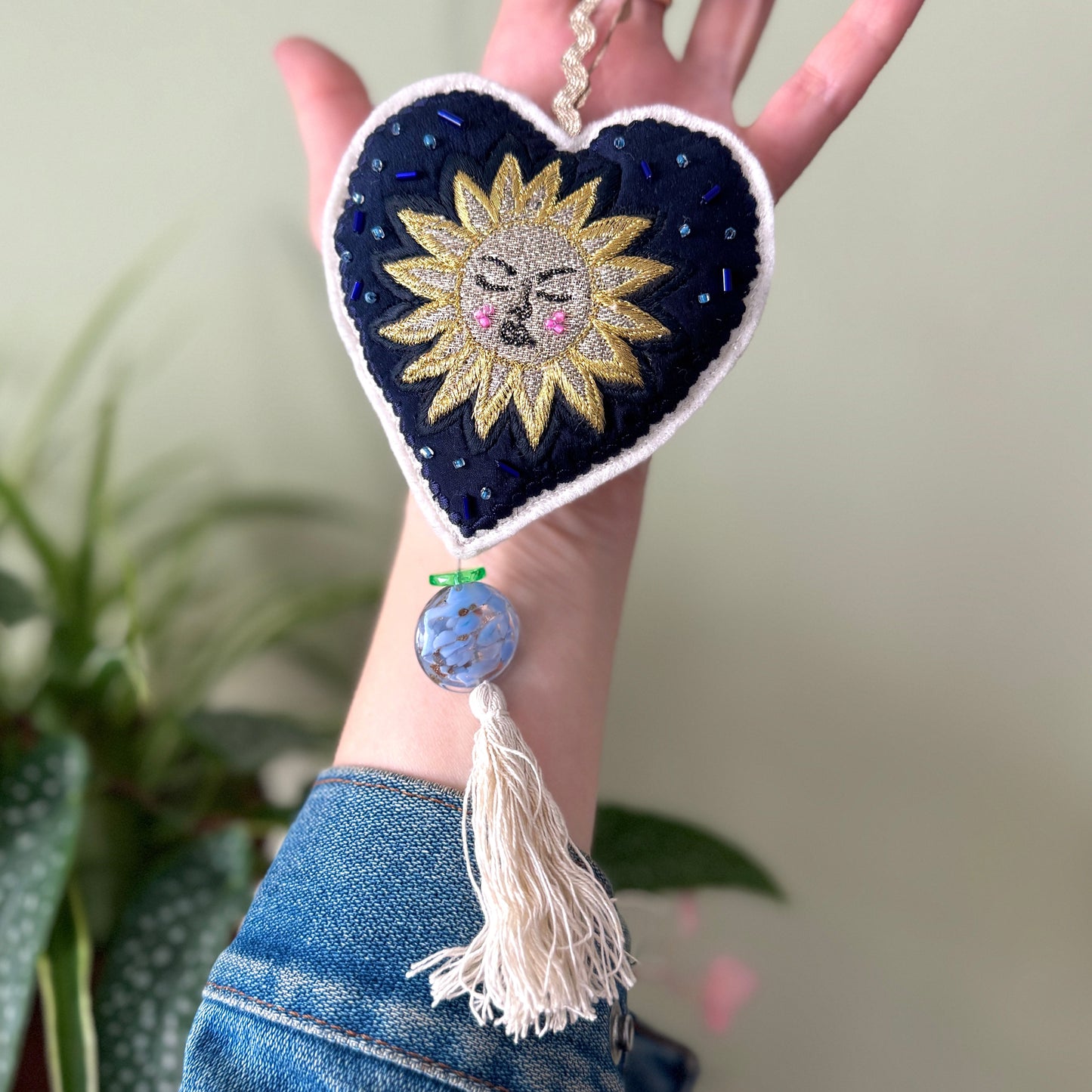 Heart-shaped decorative item with a sun design held by a hand against a blurred plant background