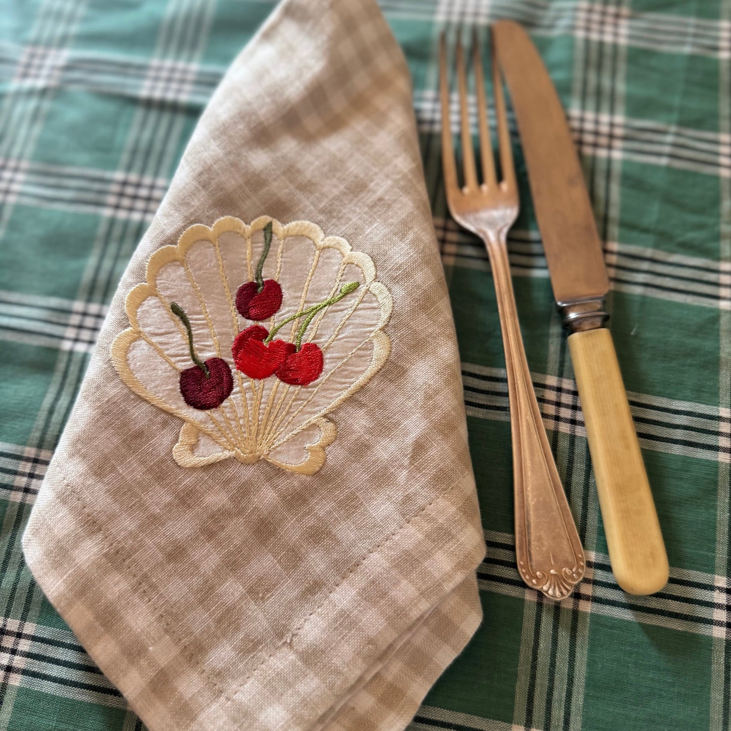Napkin with cherry design on a plaid tablecloth with fork and knife.