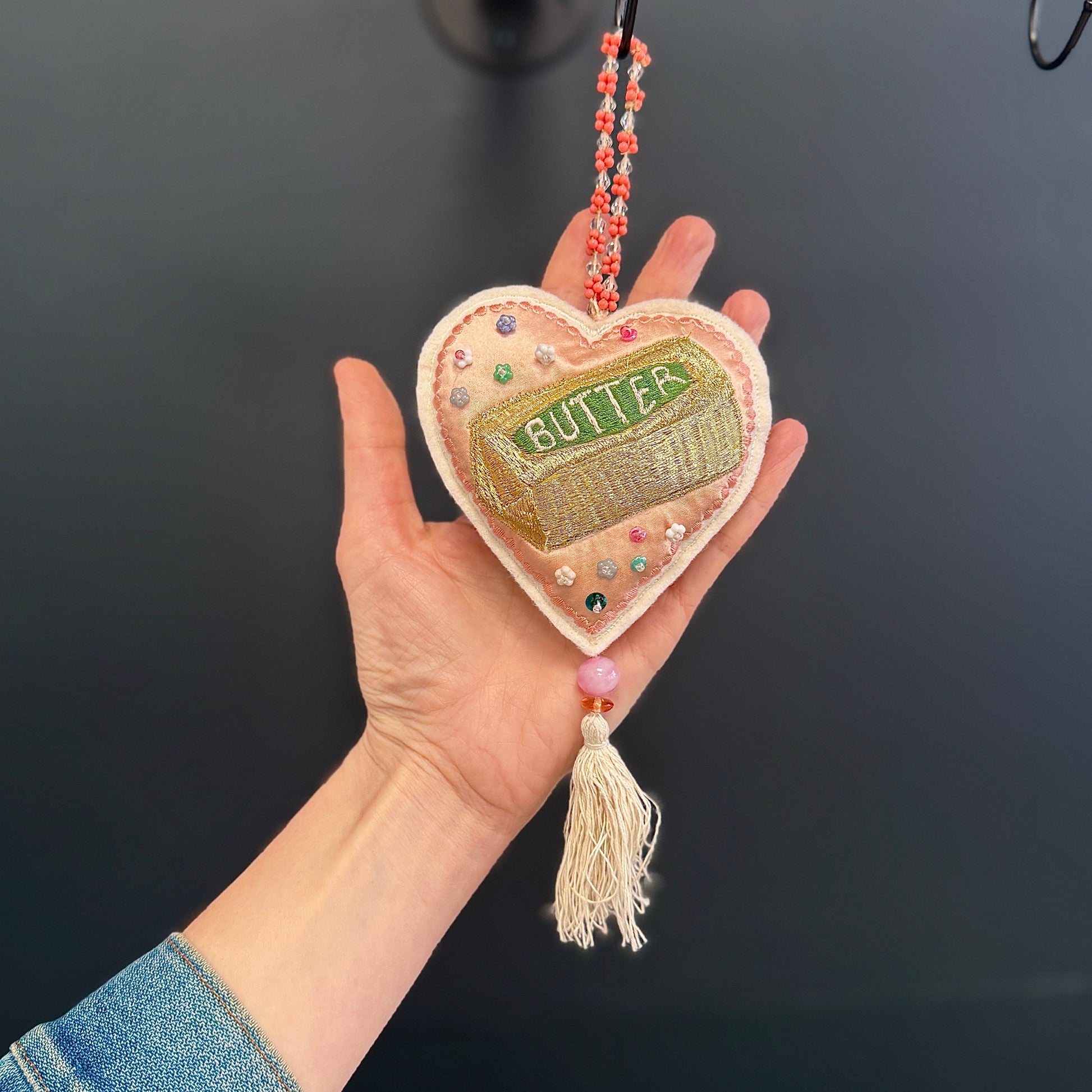Heart-shaped embroidered pendant with 'BUTTER' text held in a hand against a dark background