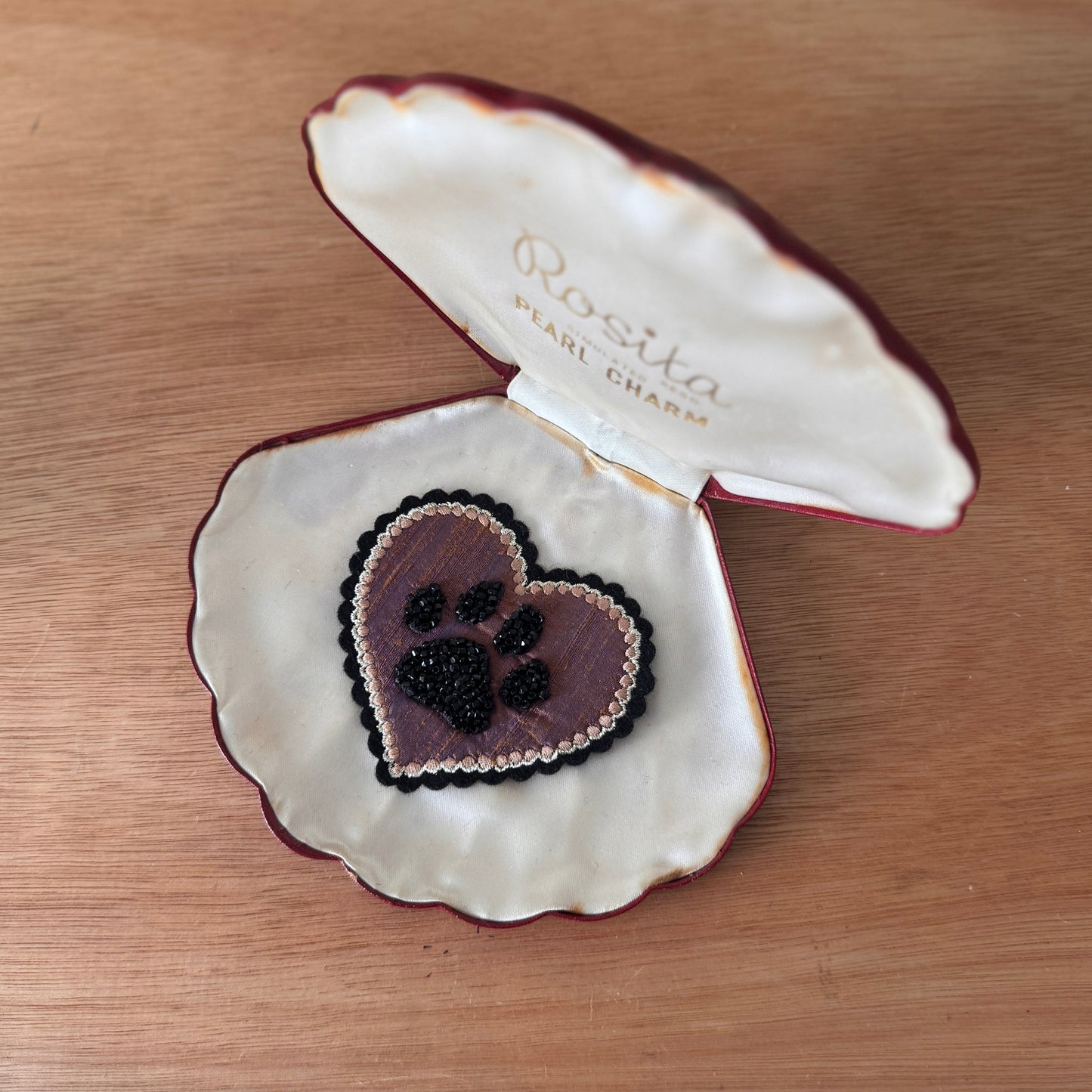 Open shell jewelry box with a heart-shaped beaded pet paw patch inside, placed on a wooden surface