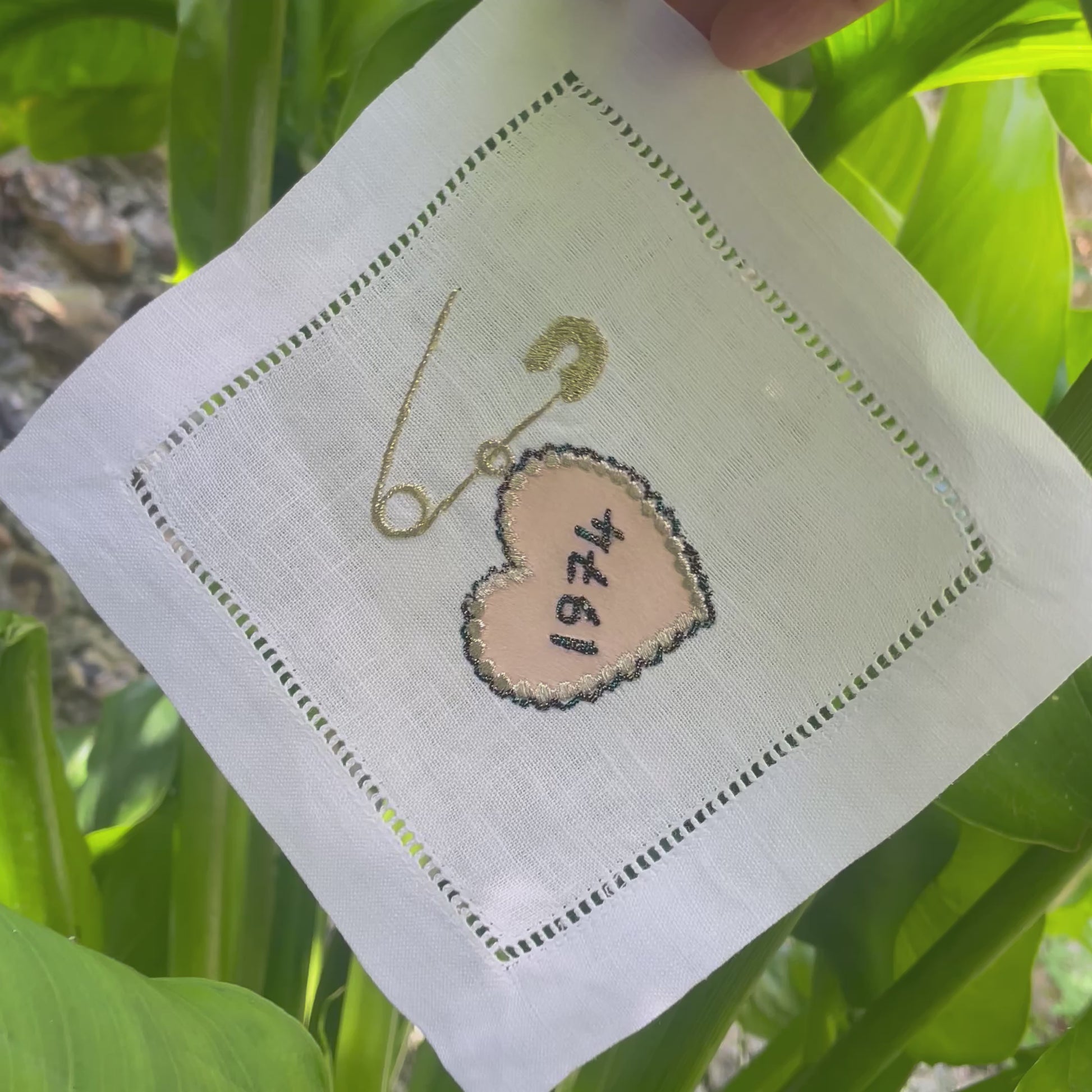 Video shows a hand holding the corner of the heart pin embroidered artwork in front of a green leafy plant. The artwork is being tilted from side to side.