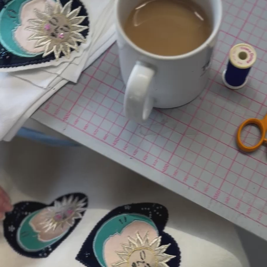 Video shows a close up of a desk edge with hands visible hand sewing small blue beads on the cosmic patches