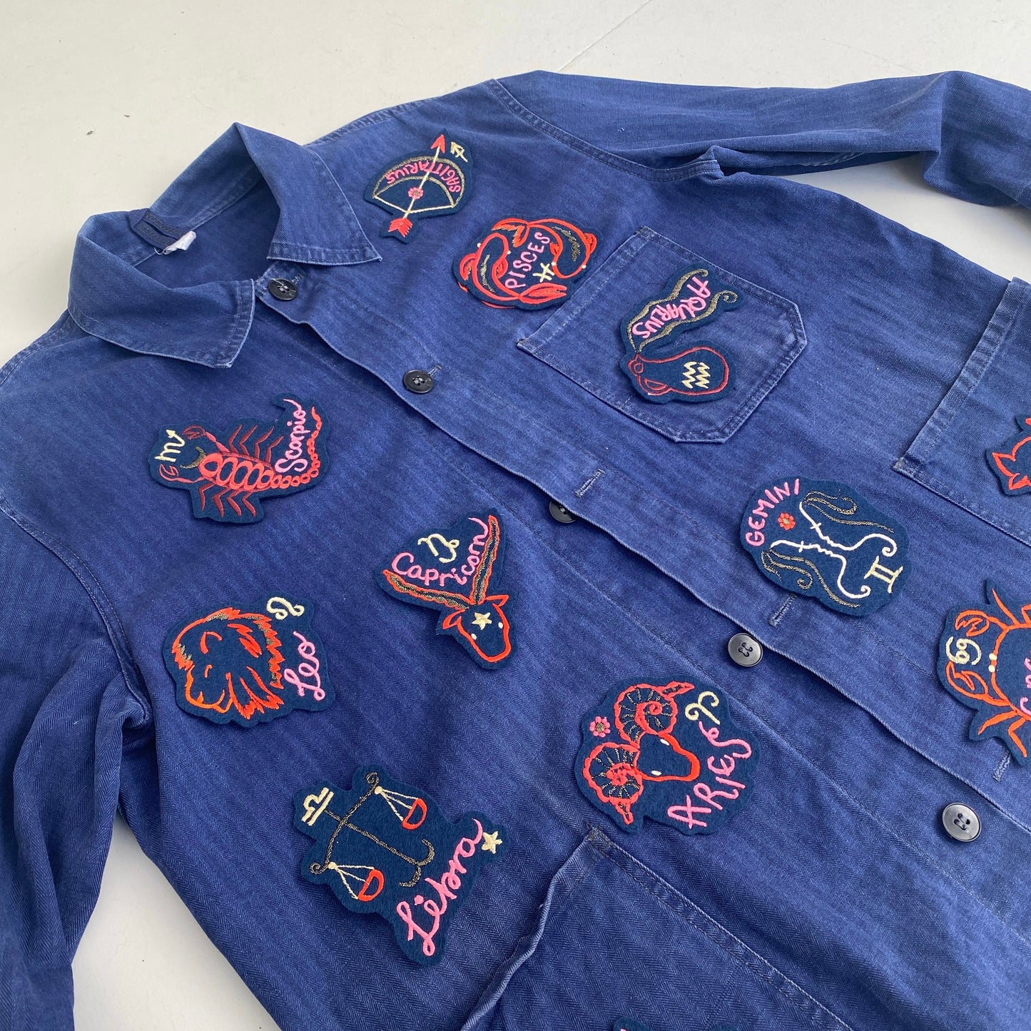 Blue 'workman's' jacket layed out and displayed with zodiac patches applied to front of jacket