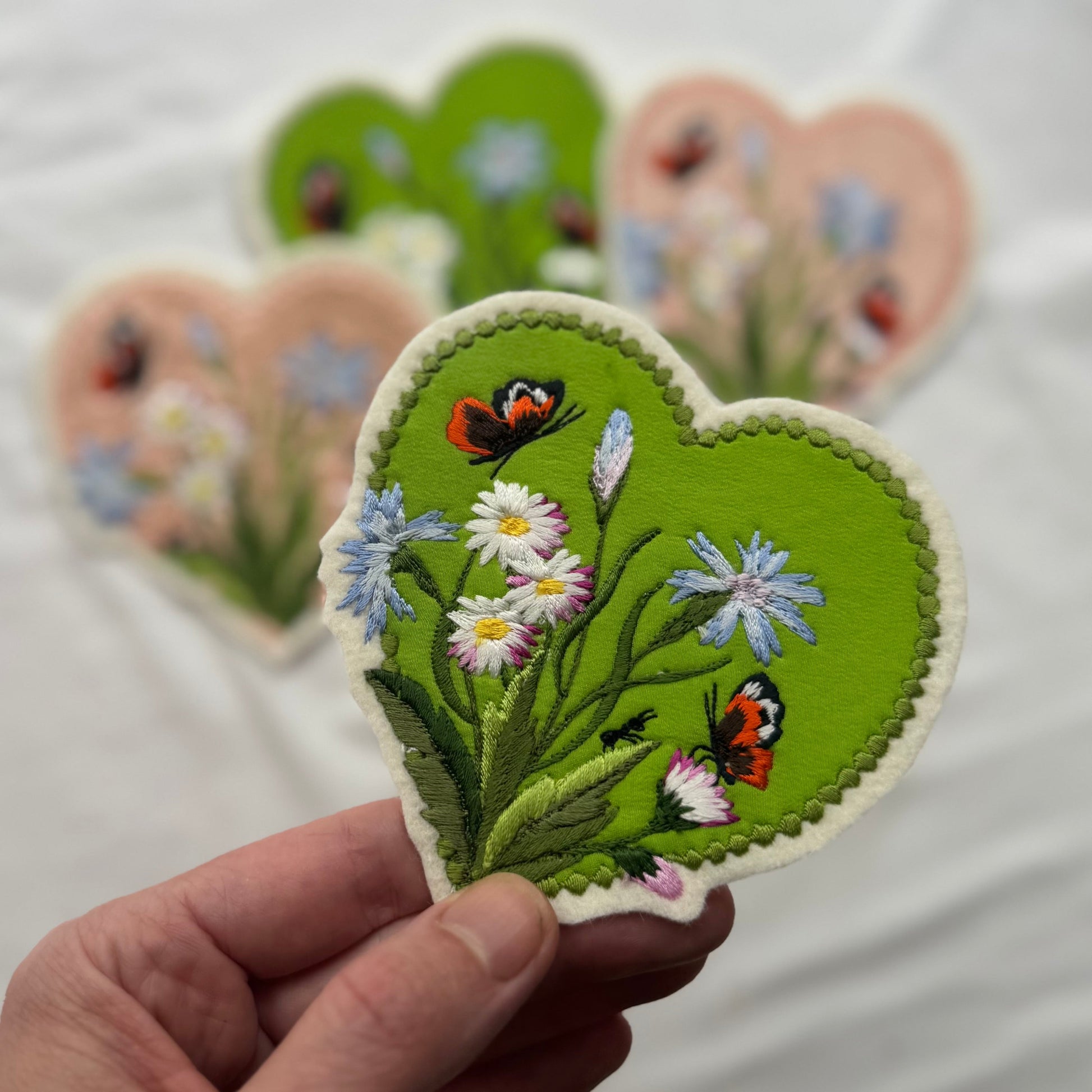 Heart-shaped embroidered patch with flowers and butterflies held by a hand against a blurred background of similar patches.
