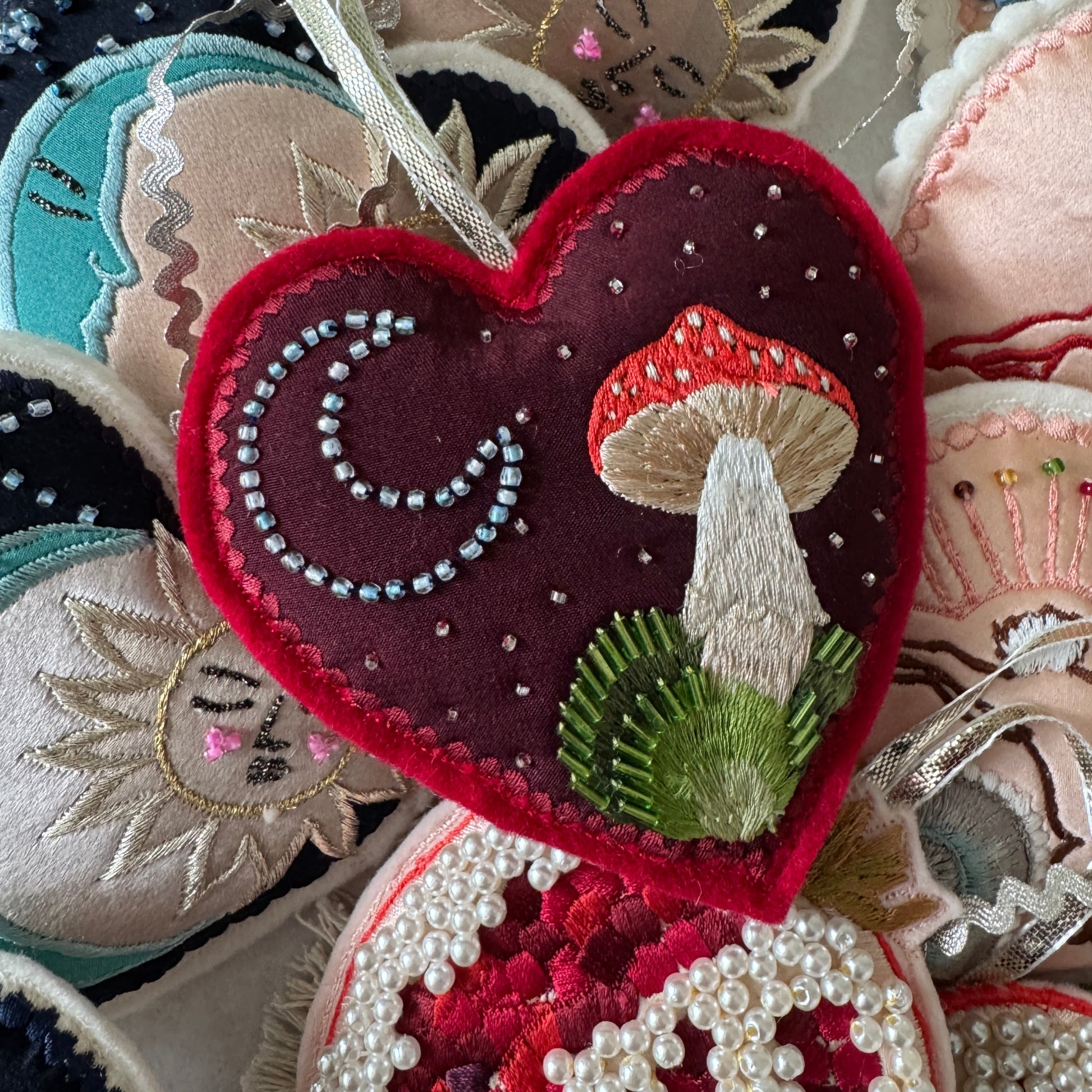 Heart-shaped embroidered ornament with mushrooms and beads, surrounded by other embroidered pieces.