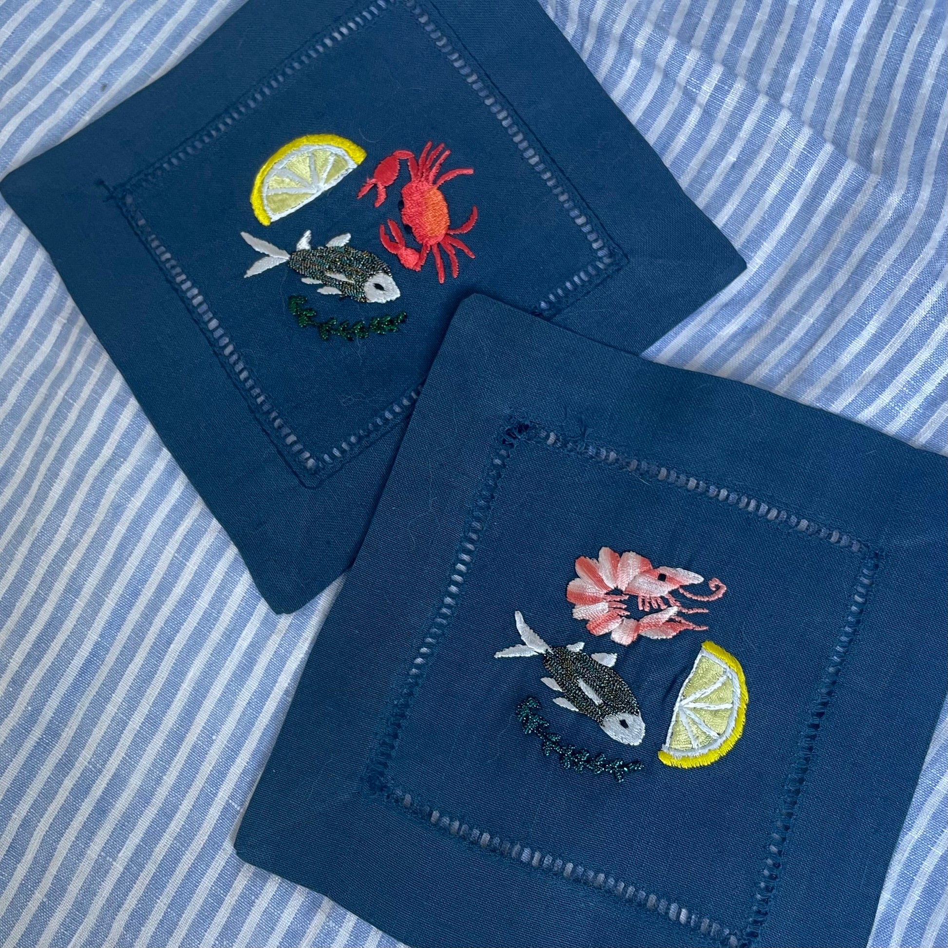 Navy blue linen cocktail napkins with sea creatures embroidery, displayed slightly overlapping on a blue and white striped cloth