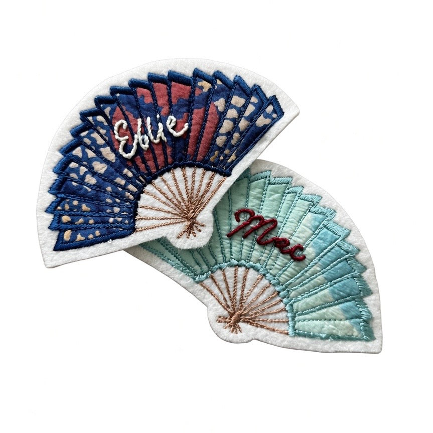Two embroidered patch designs shaped like folding fans, shown overlapping on a plain white background. One patch has blue, red, and white fan sections with the name 'Ellie' in white script.