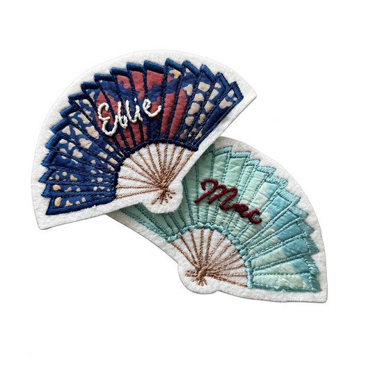Two embroidered patch designs shaped like folding fans, shown overlapping on a plain white background. One patch has blue, red, and white fan sections with the name 'Ellie' in white script.