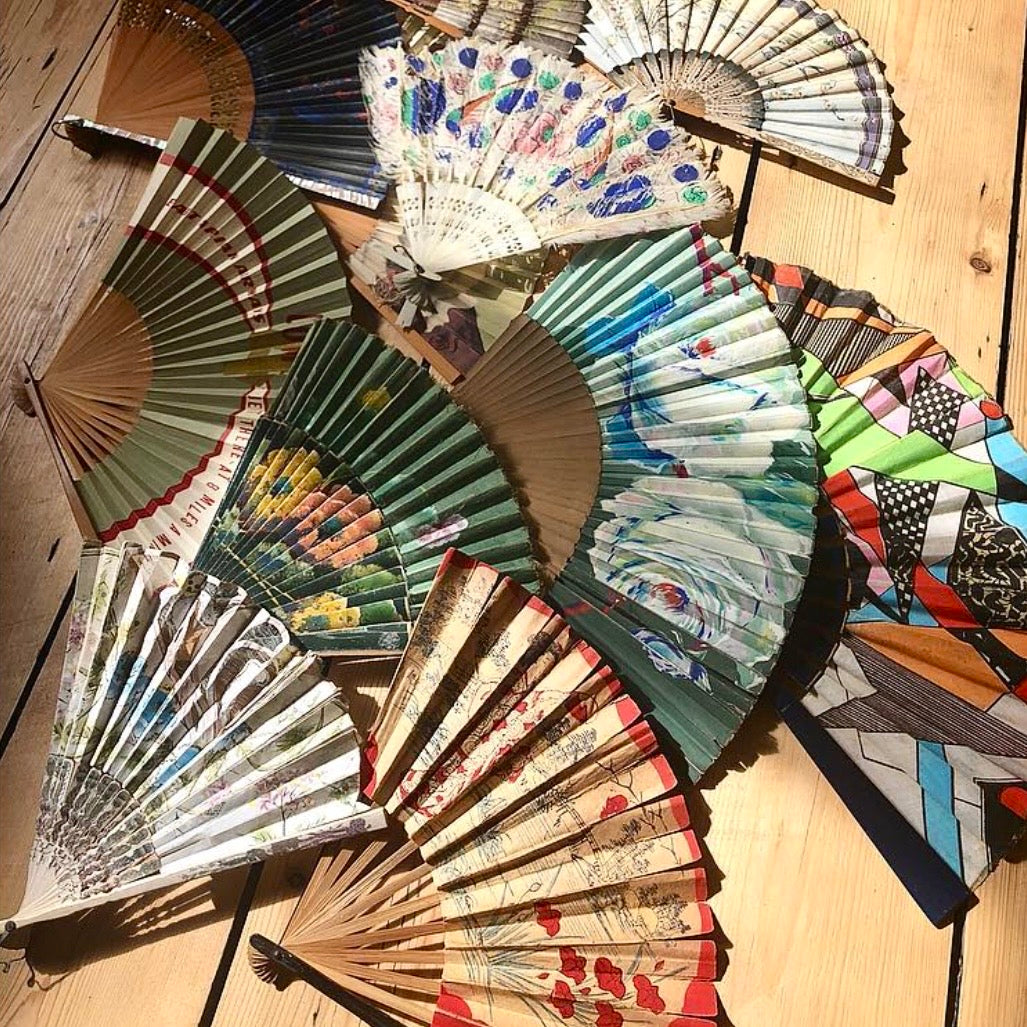 A collection of traditional folding hand fans with various printed designs, including floral, landscape, and geometric patterns, arranged on a light wooden surface.