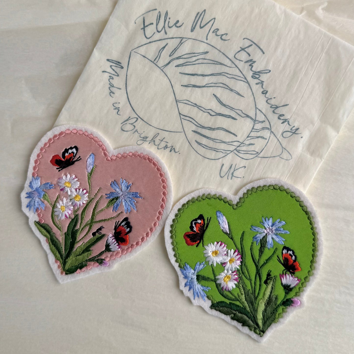 Two embroidered heart-shaped patches with flowers and butterflies on a white fabric background with 'Ellie Mac Embroidery' branding.