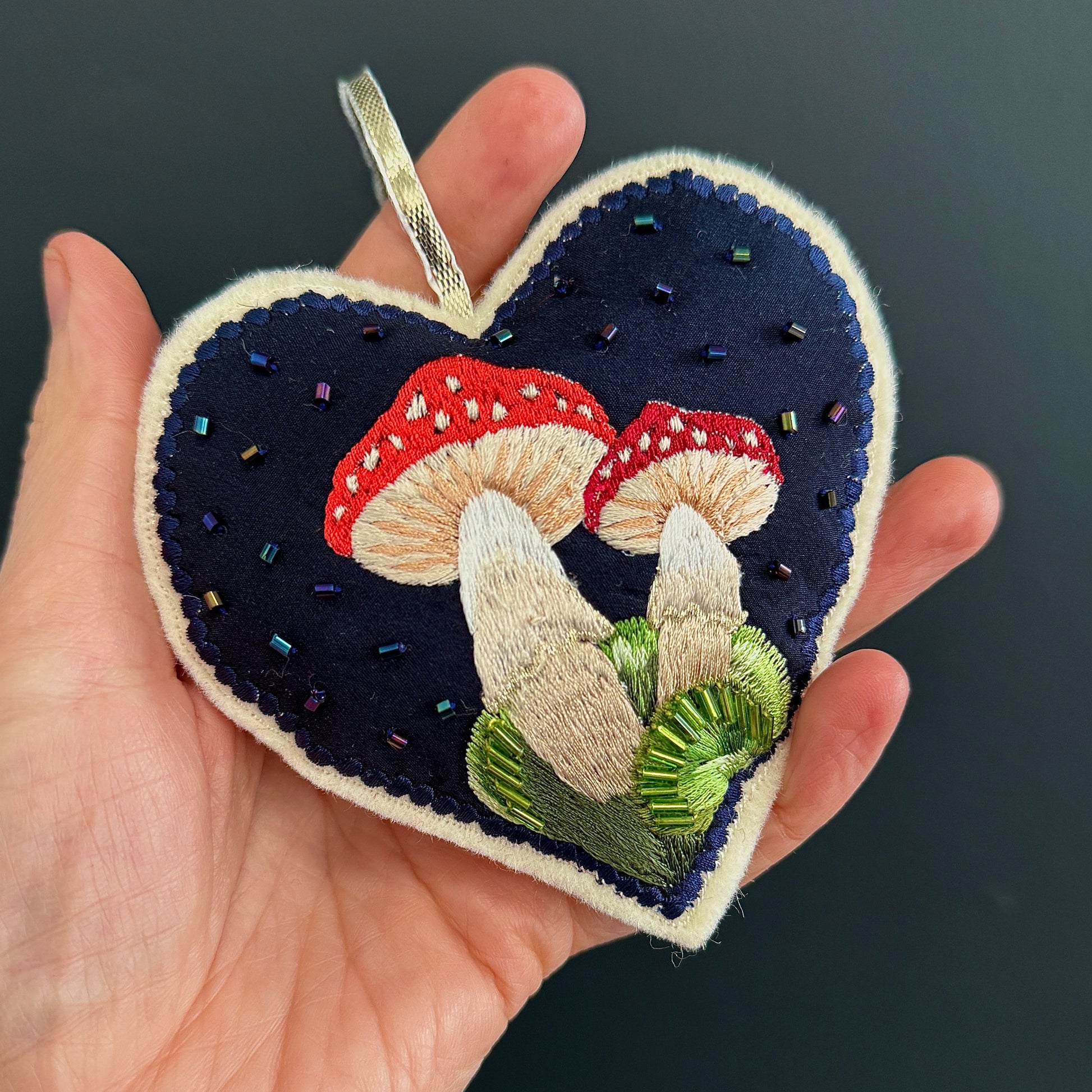 Heart-shaped embroidered patch with mushrooms held in a hand against a dark background