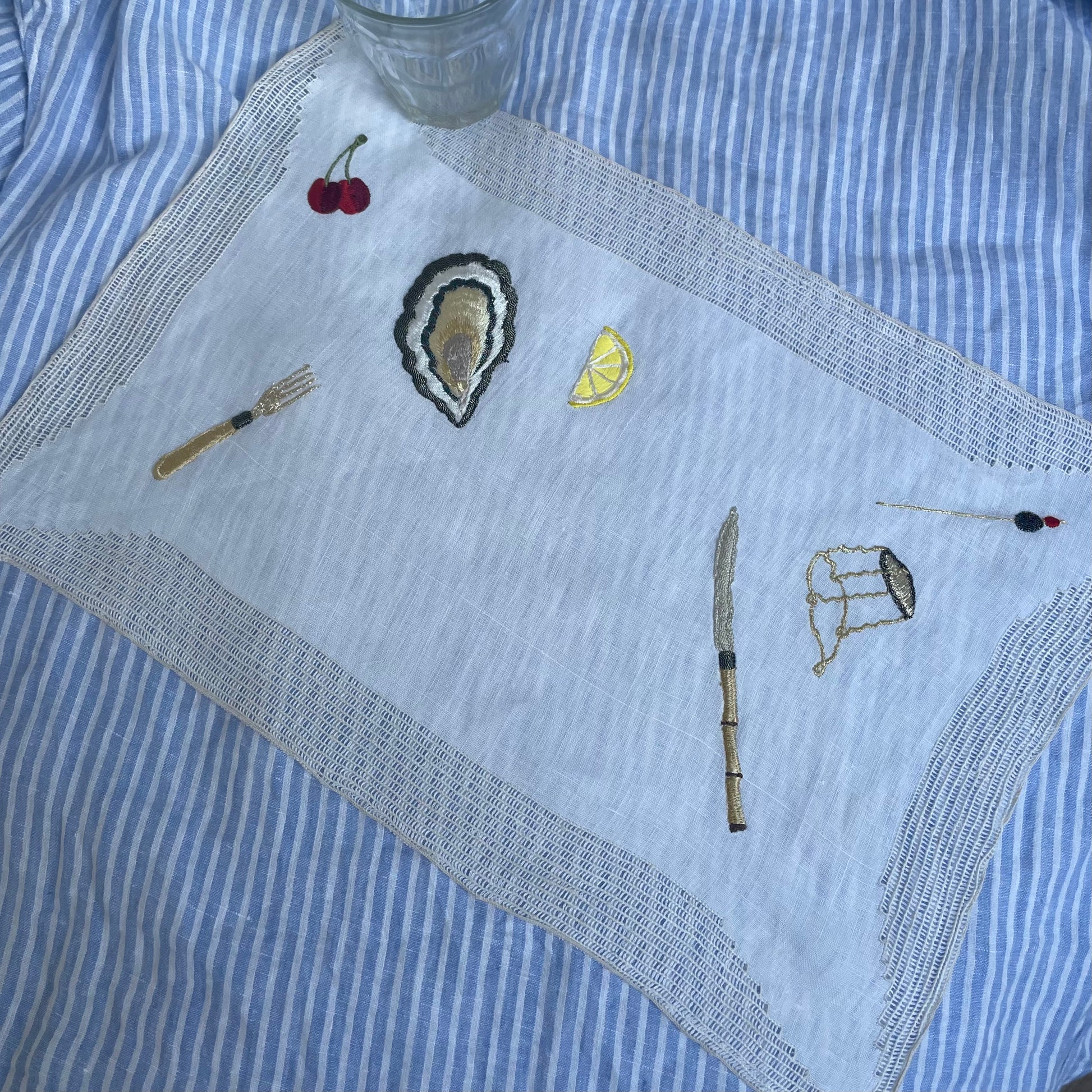 Vintage linen placemat with cherry, oyster, fork, knife, lemon wedge and champagne cork embroidery, displayed on a white and blue striped cloth with a drinking glass just visible in corner