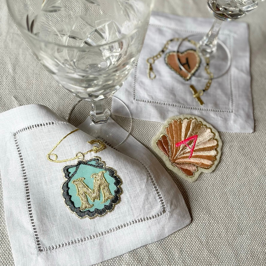 Two embroidered artworks and an embroidered shell patch on a linen table cloth with two glasses
