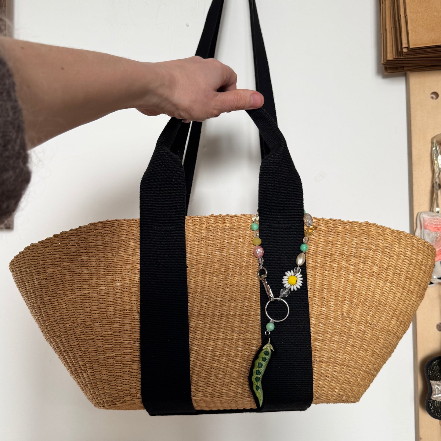 Woven bag with black straps held by a hand against a white wall.