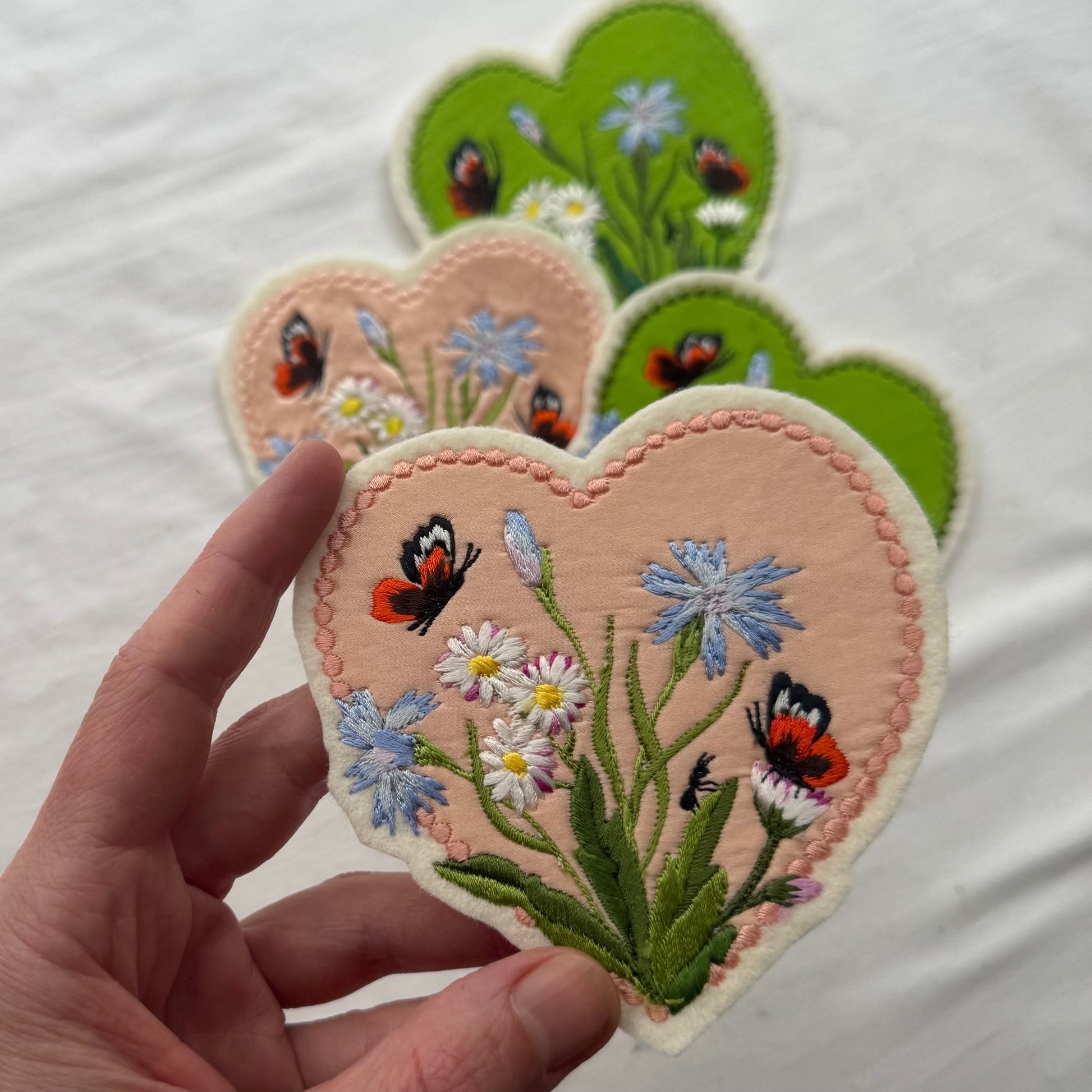 Heart-shaped embroidered patches with flowers and butterflies held by a hand on a white background
