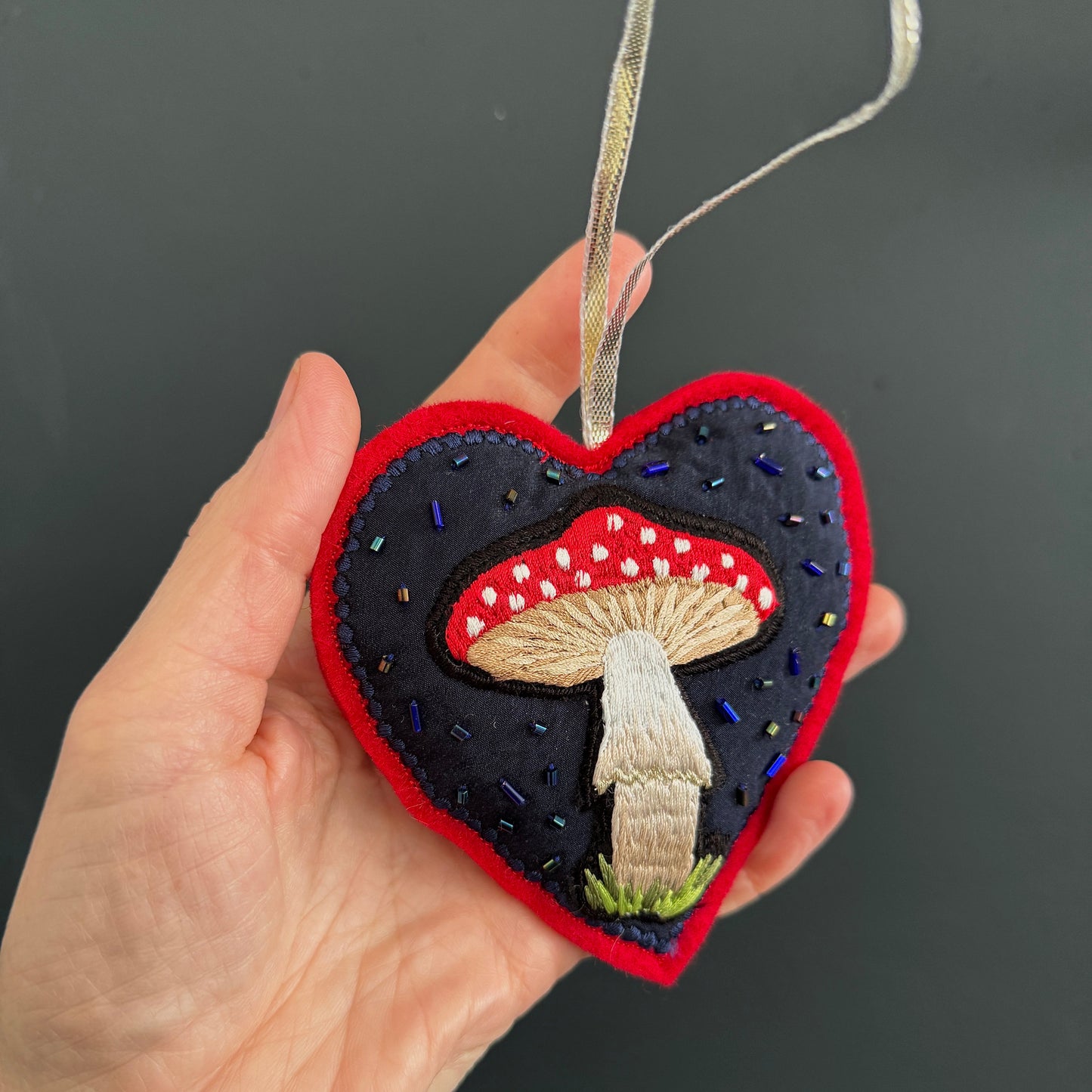 Heart-shaped embroidered ornament with mushrooms held by a hand against a dark background