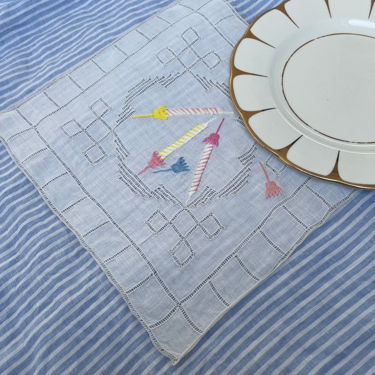 Vintage linen white cocktail napkin with embroidered birthday candles motif shown on a blue and white striped cloth with the corner of a small white and gold plate also visible