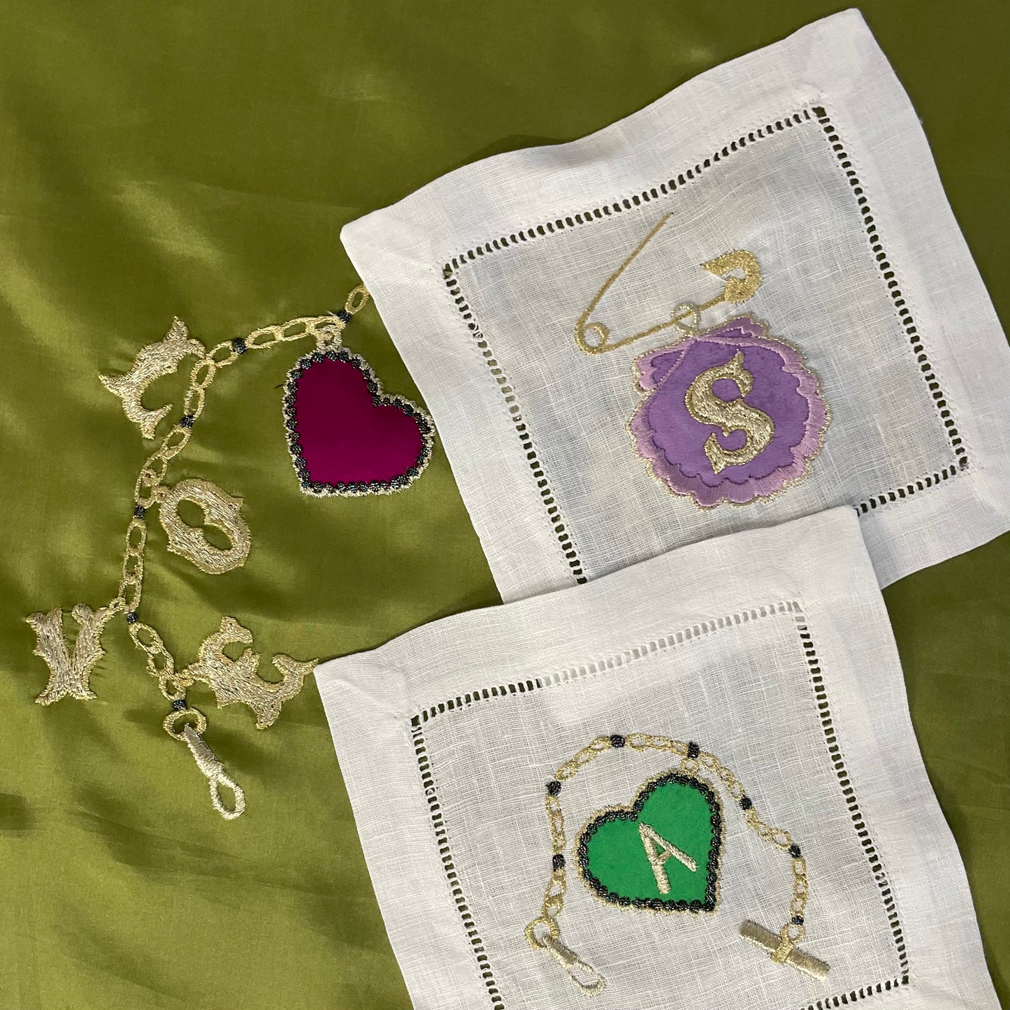 Two embroidered artworks from the Love Token collection on a piece of olive green fabric which has an embroidered charm bracelet on