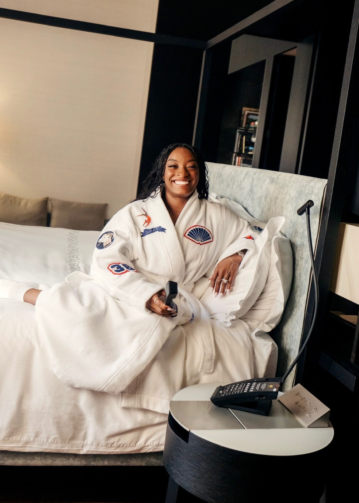 Simone Biles in collaboration with Mandarin Oriental in a robe sitting on a bed in a hotel room with a nightstand and phone.