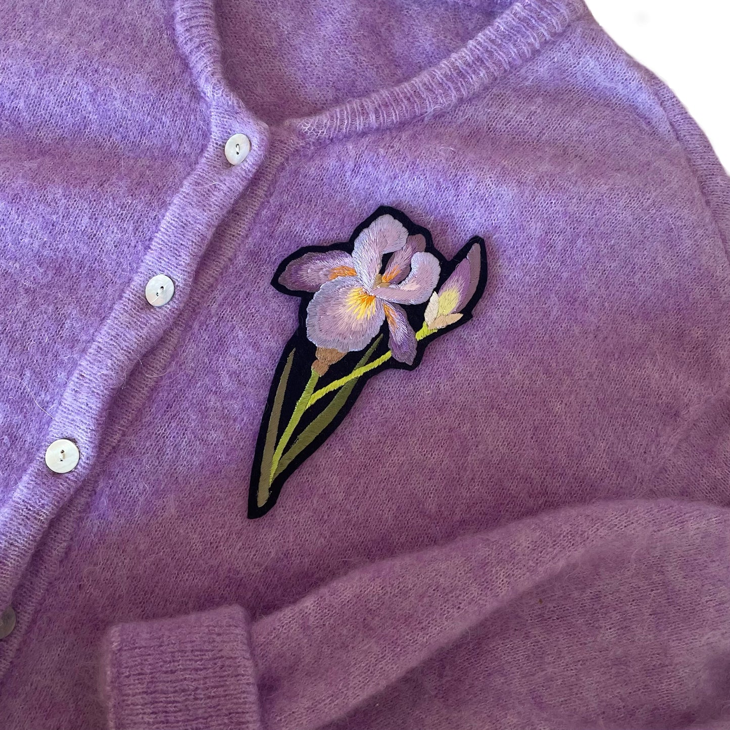 Embroidered iris patch shown on the breast of a purple cardigan