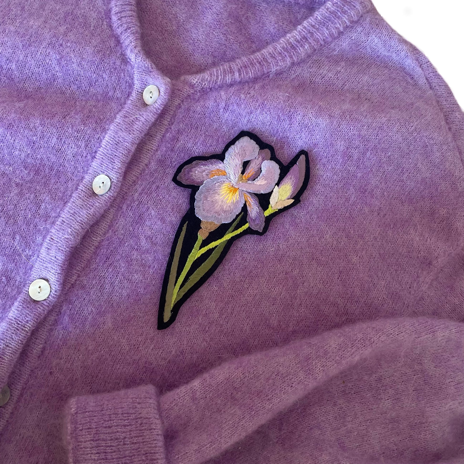 Embroidered iris patch shown on the breast of a purple cardigan