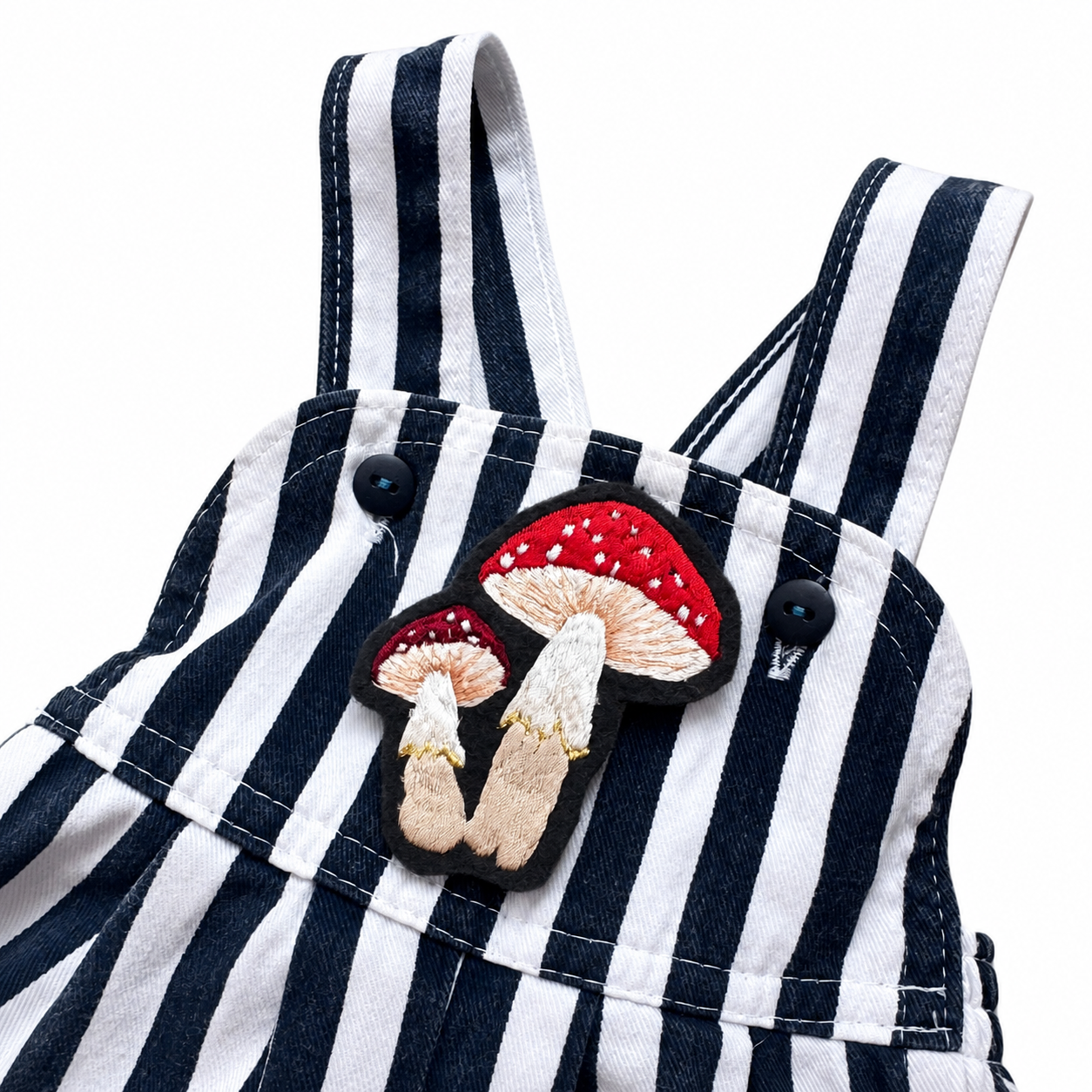 Close-up of a navy and white striped baby overalls garment with a decorative embroidered mushroom patch on the chest pocket area.