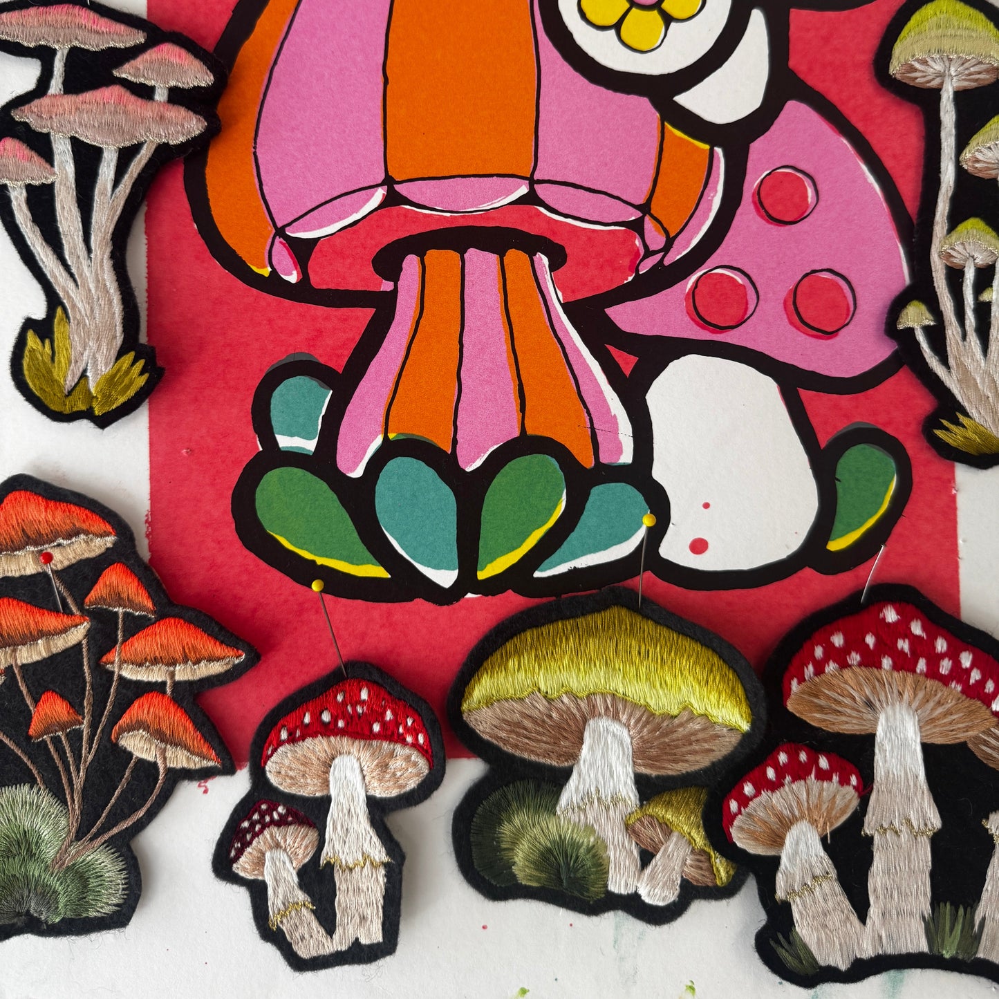 A close-up of several embroidered mushroom patches arranged on a red background. The patches feature different mushroom designs.
