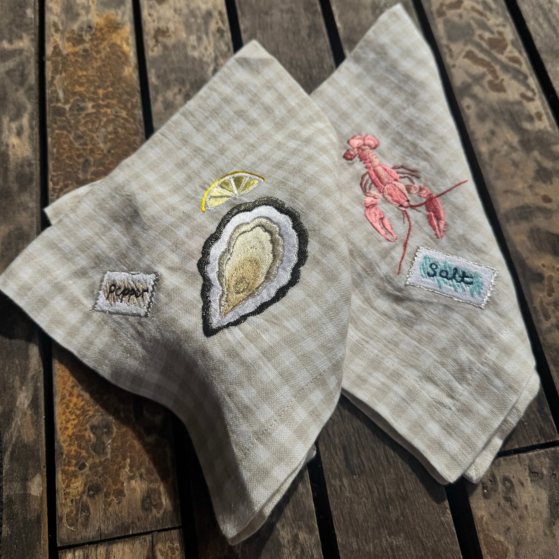 Two embroidered towels on a wooden surface, one with an oyster and lemon design and the other with a lobster design.