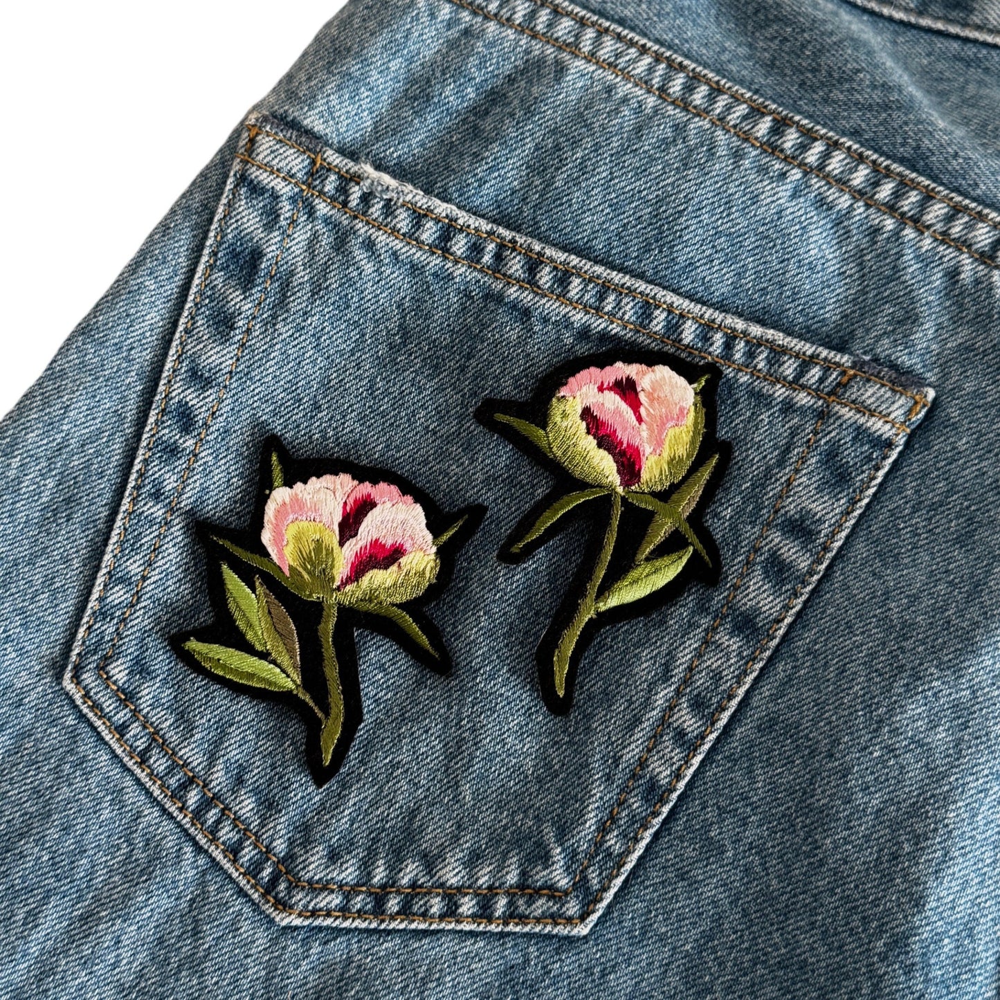 Two embroidered floral patches with pink and white flowers and green leaves, placed on a blue denim pocket.