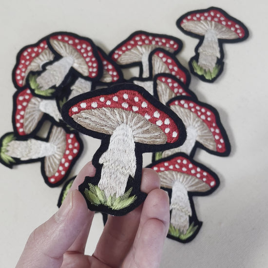 Video of a hand holding an embroidered mushroom patch, holding it close to the camera and tilting left and right. More mushroom patches are visible in the background