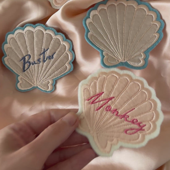 Video of a hand holding up one of four different silk applique shell patches to show the embroidered details. Other shell patches are placed on pink silk fabric