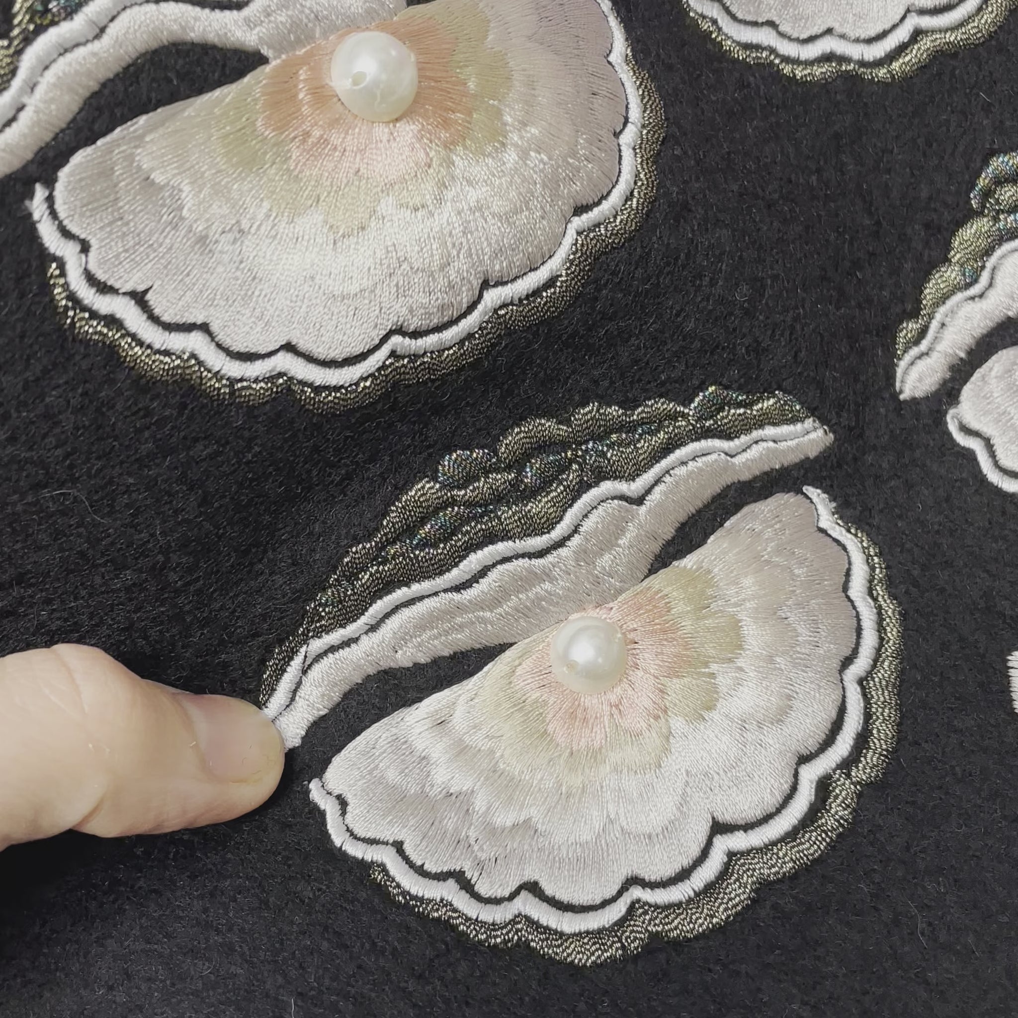 Close-up video of pre-cut oyster with pearl embroidered patches. A hand is just seen tilting the embroidery to catch the light and show the embroidery