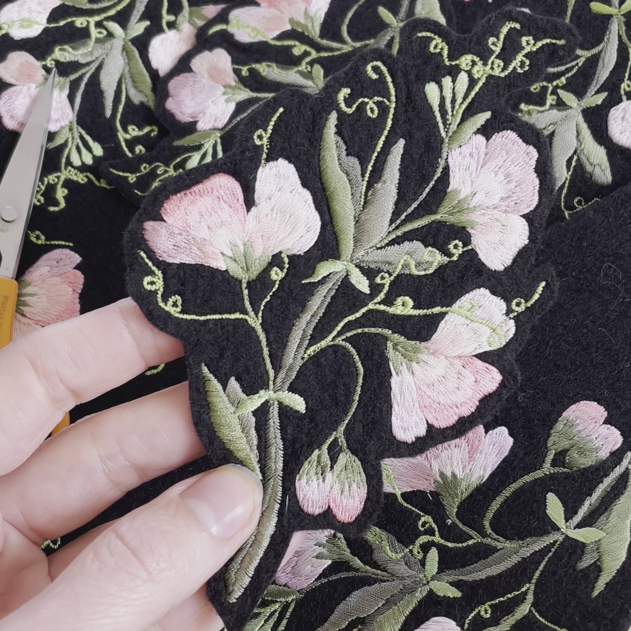 Video of a hand holding a sweet pea embroidrered patch and tilting it from side to side to show the embroidery. More sweet pea patches can be seen in the background