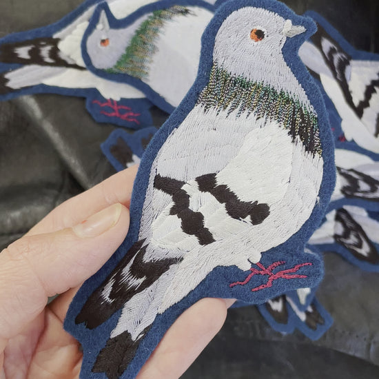 Video of a pigeon embroidered patch held in someone's hand who is tilting the patch at the camera. A stack of further pigeon embroidered patches are visible in the background