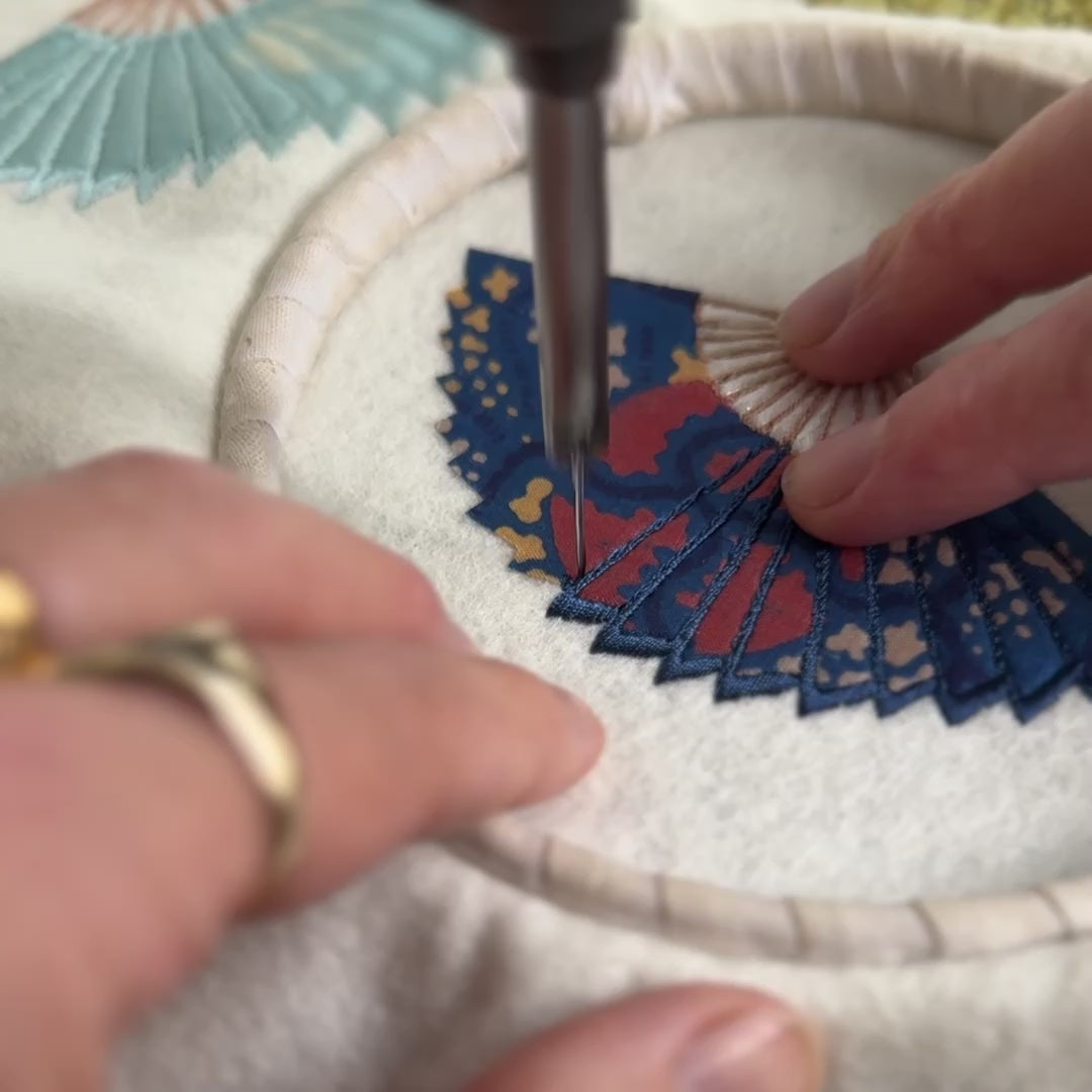Close-up of needle on a freehand embroidery machine stitching navy thread on a silk fan design on cream fabric. The fabric is in a small hoop with the fingertips of two hands shown guiding the hoop and fabric