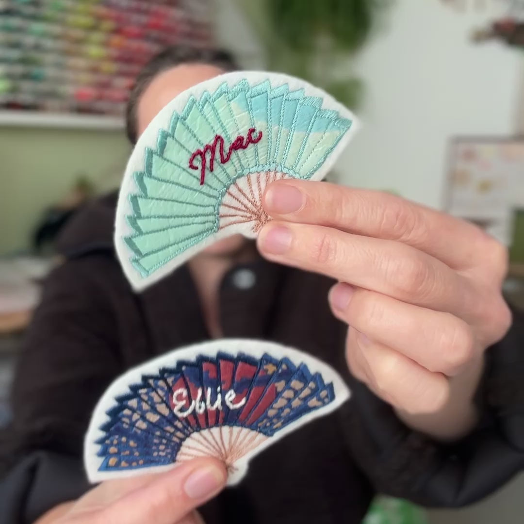 Ellie Mac holding up two fan patches and tilting them left and right to show details. Sat in her embroidery studio, wearing a brown jacket