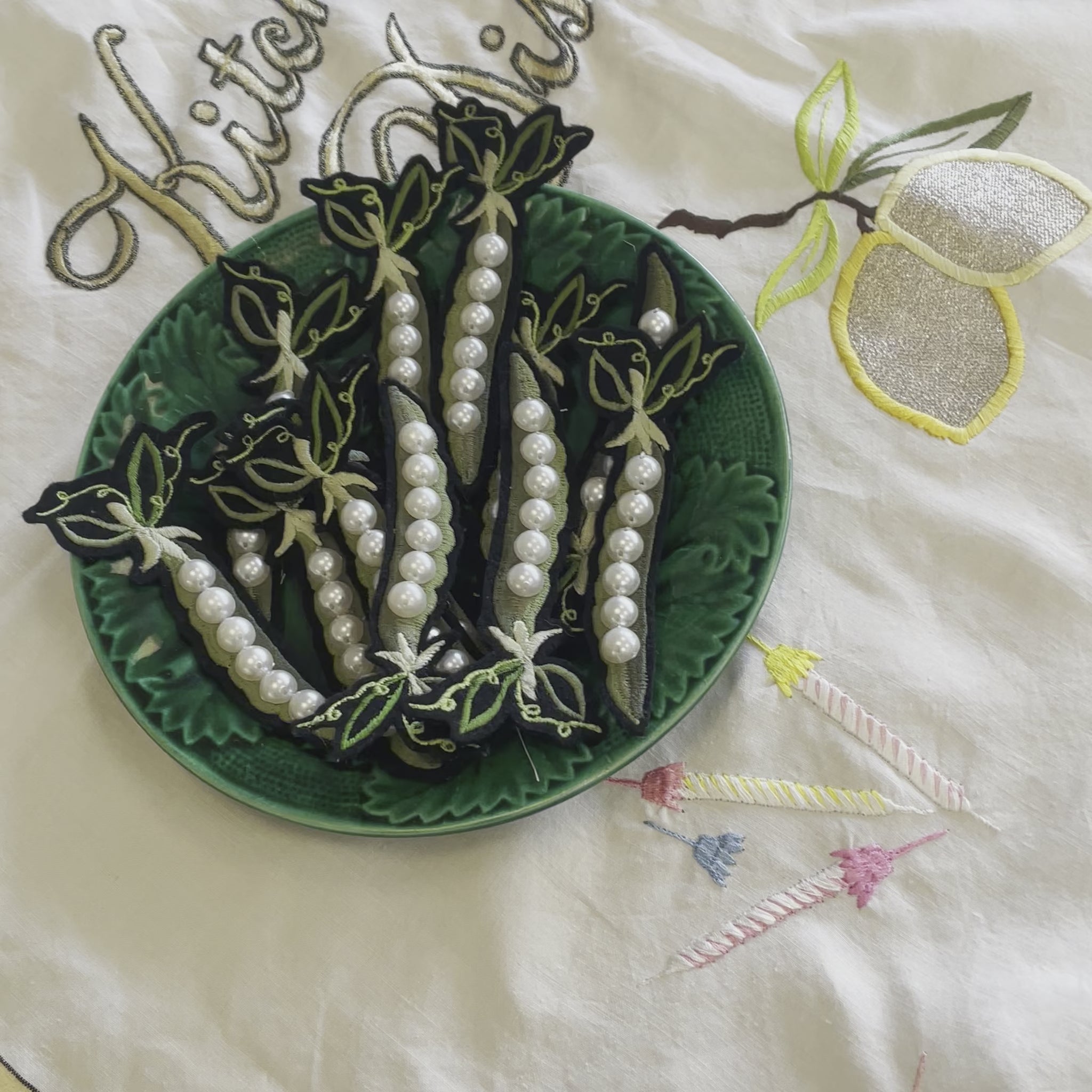 Video shows a green bowl with a number of pea and pearl embroidered patches on an embroidered table cloth. A hand picks up one of the pea patches and holds it closer to the camera
