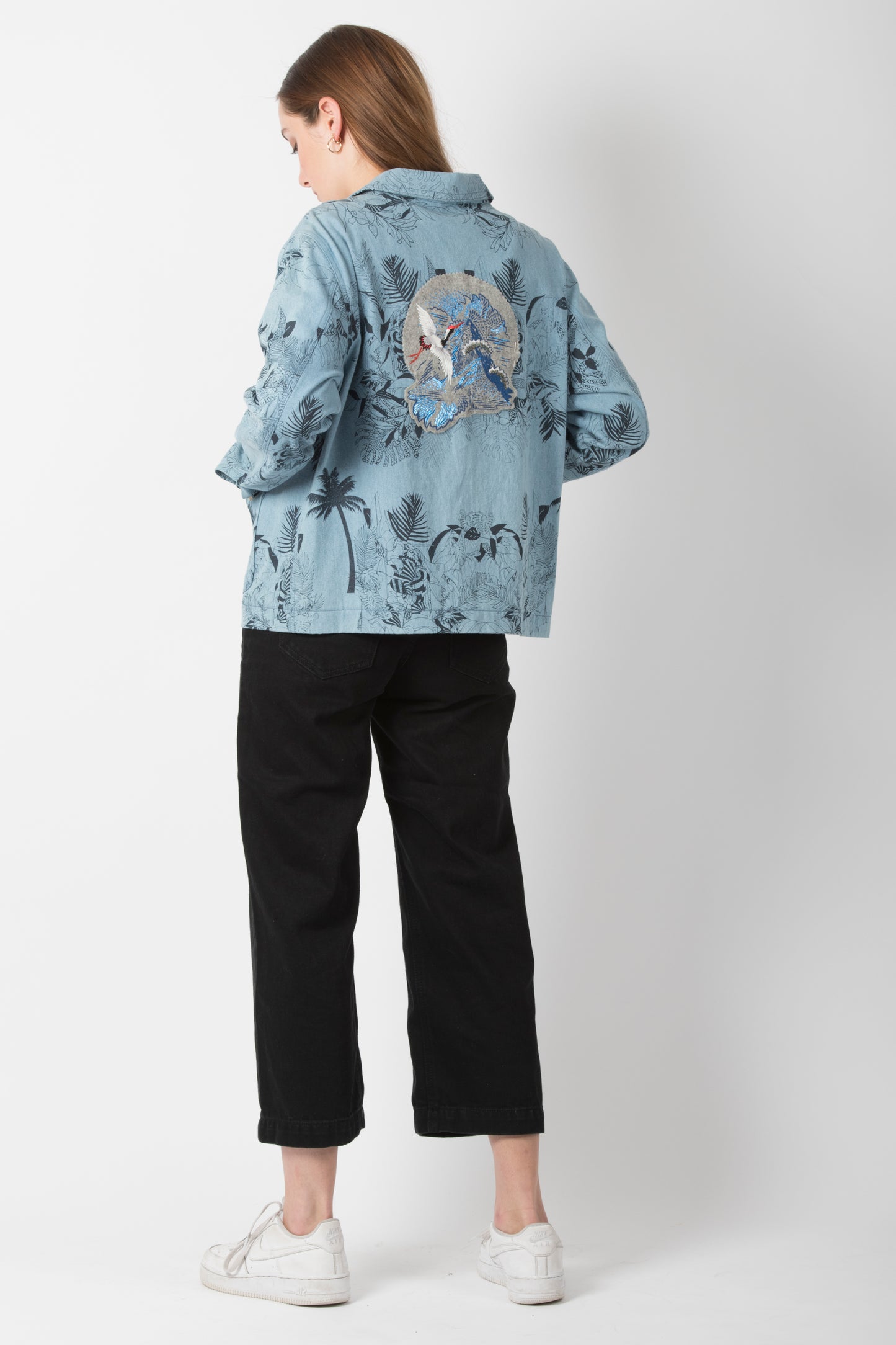 The back of a woman is seen, she is wearing a pale blue denim jacket, black trousers and white trainers. On the back of the denim jacket she is wearing is the foil printed & embroidered mountain crane patch