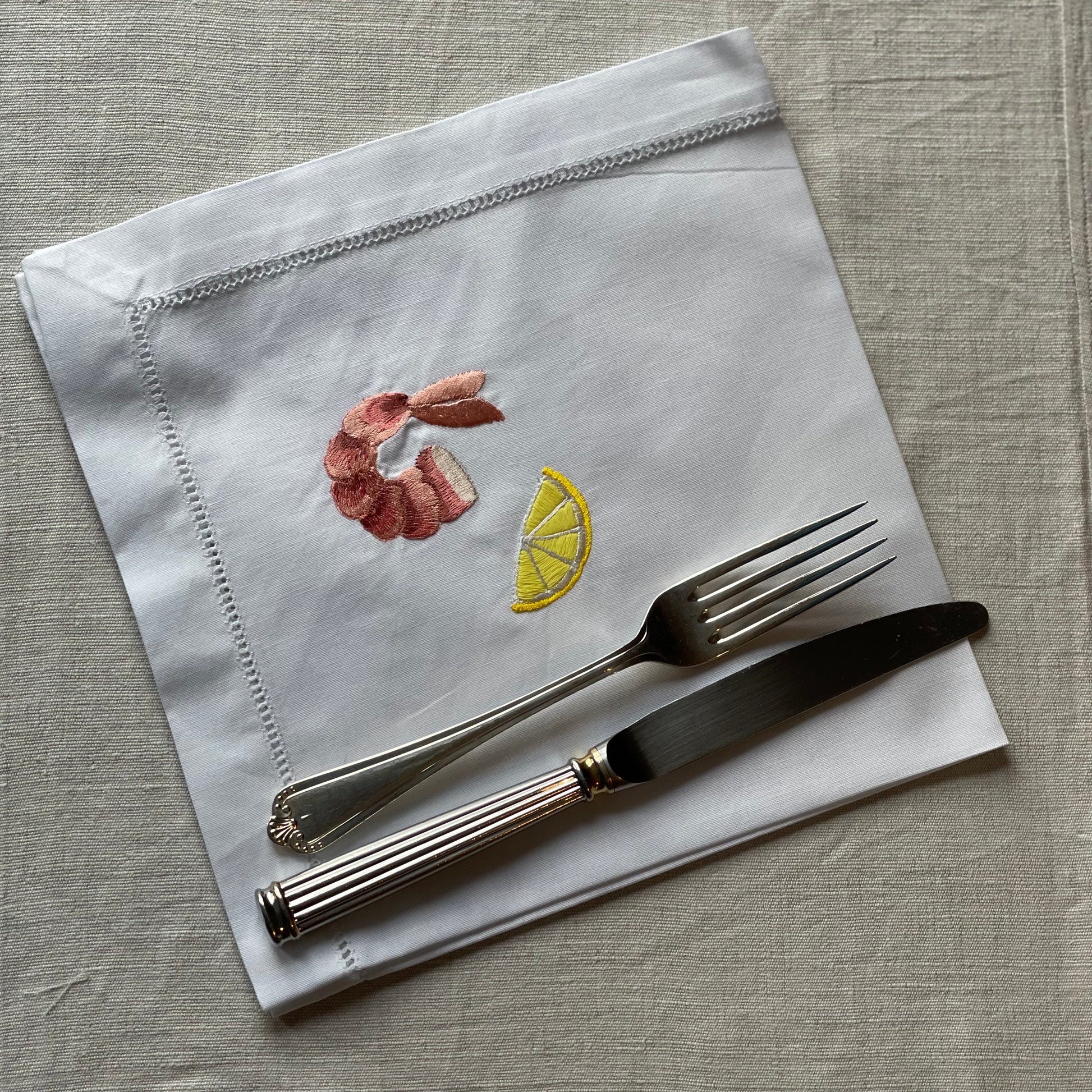 White napkin with embroidered prawn and lemon on and a metal knife and fork laid on top