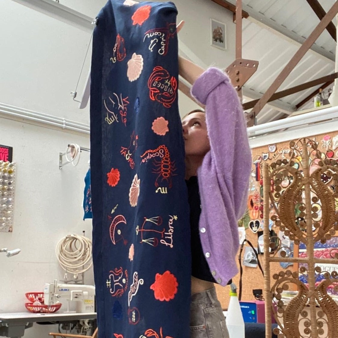 Person in lilac jumper standing behind and zodiac embroidered art work banner and looking at the back of it. The banner is hanging up, vertically, in Ellie Mac's studio