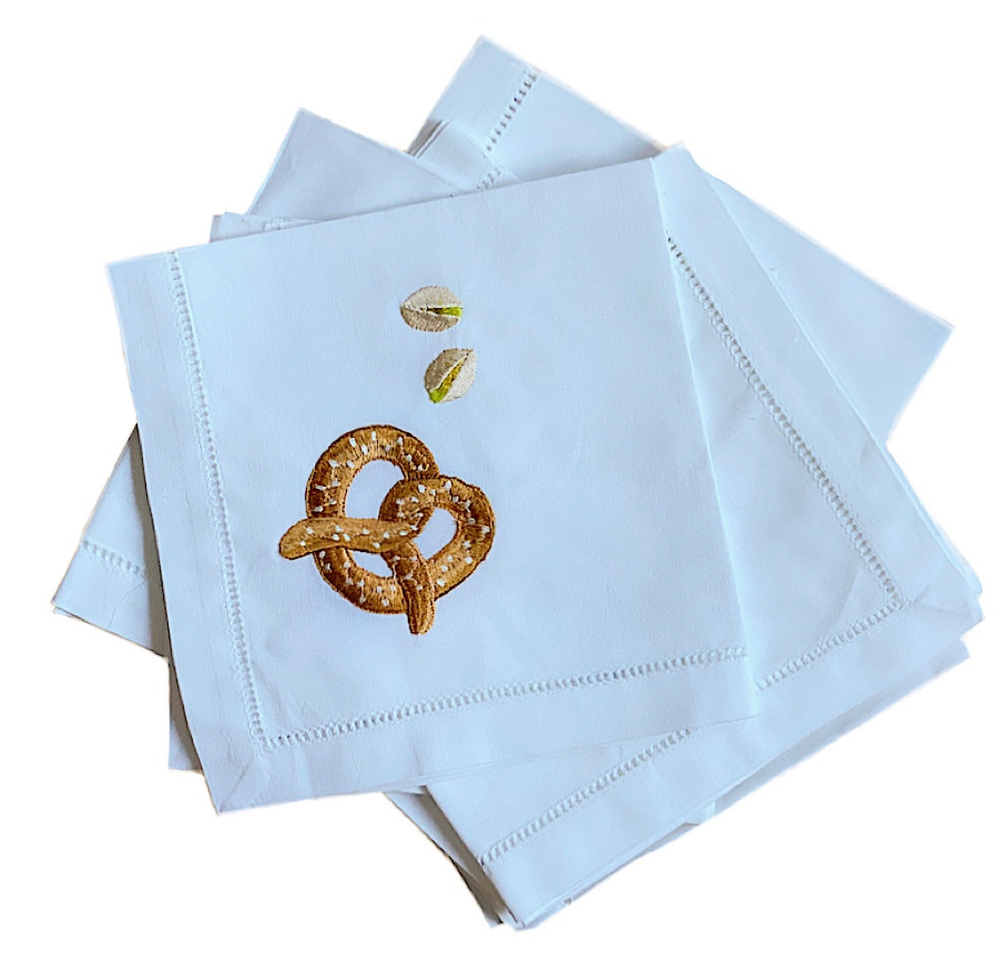 Pile of white napkins. Top napkin features embroidered pretzel and two embroidered pistachios